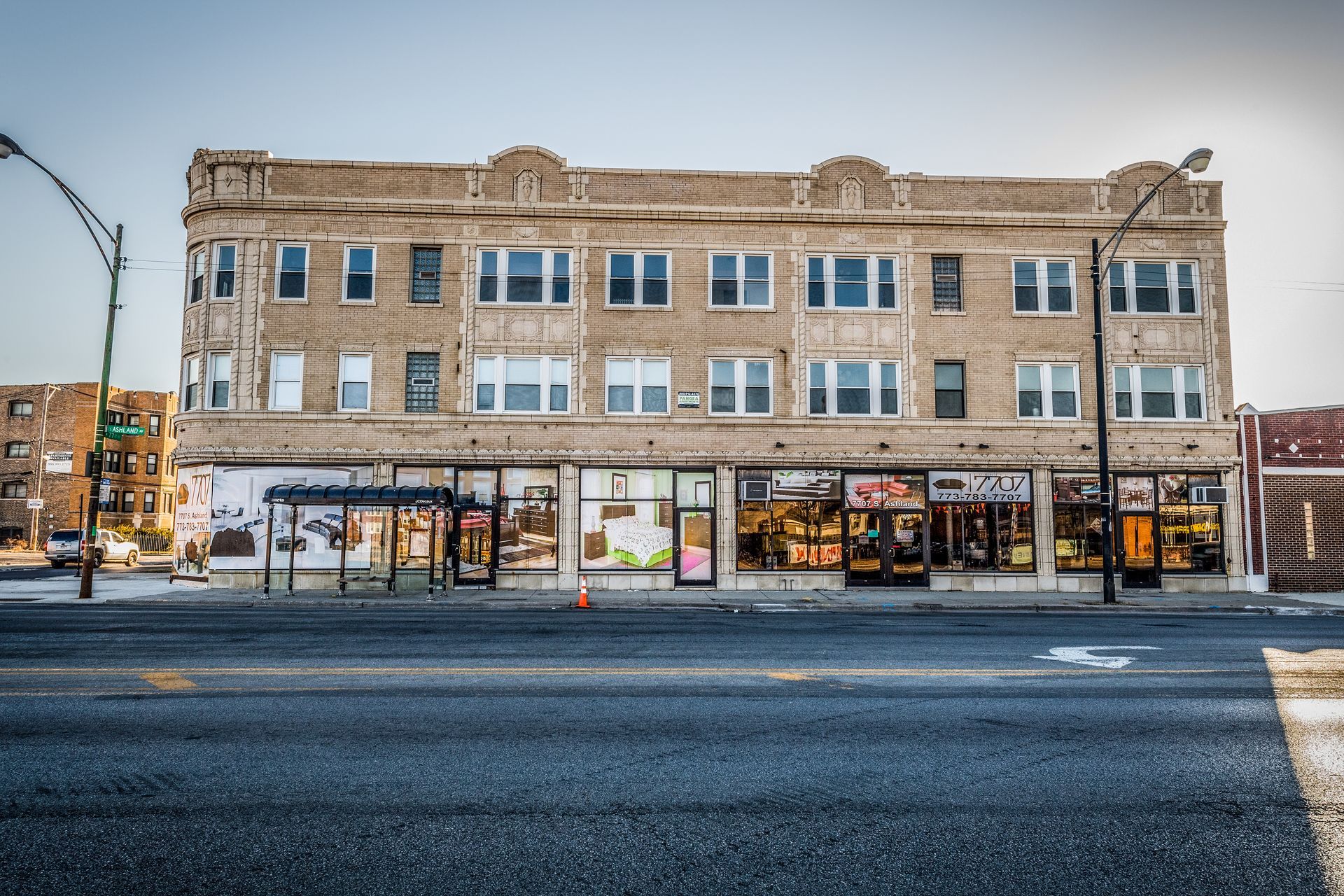 A three-story brick building with storefronts and a bus stop along a street.