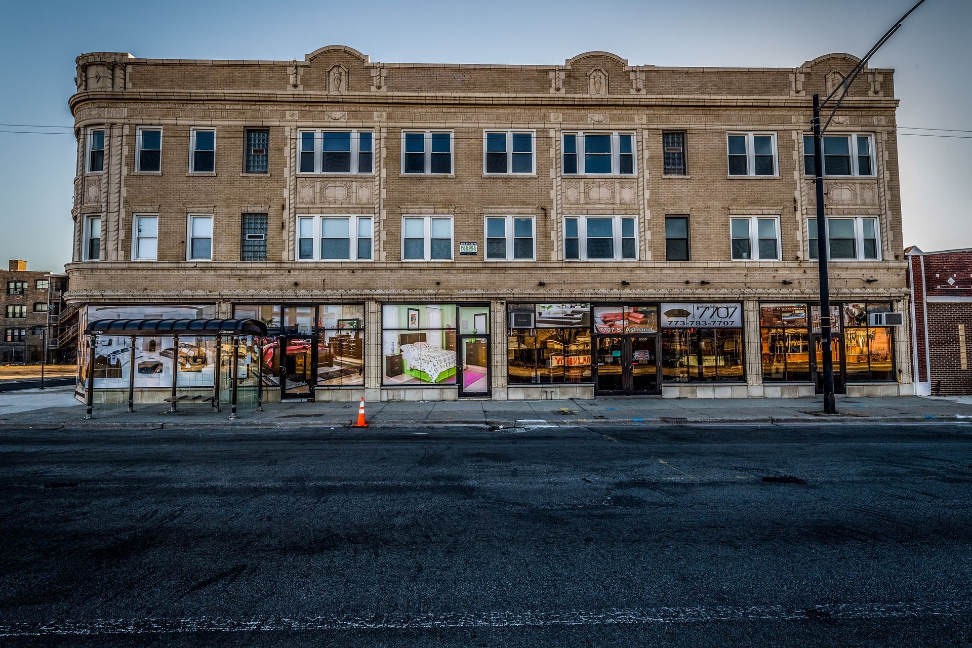 Three-story brick building with storefronts on the first floor, bus stop, and cracked asphalt road.