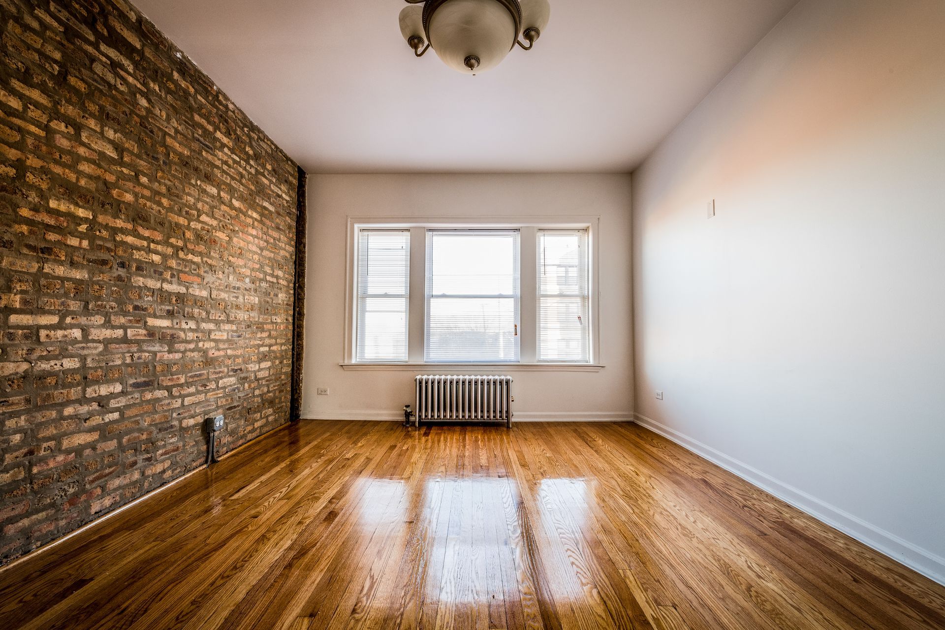 Empty room with exposed brick wall, window, hardwood floors, and overhead light.