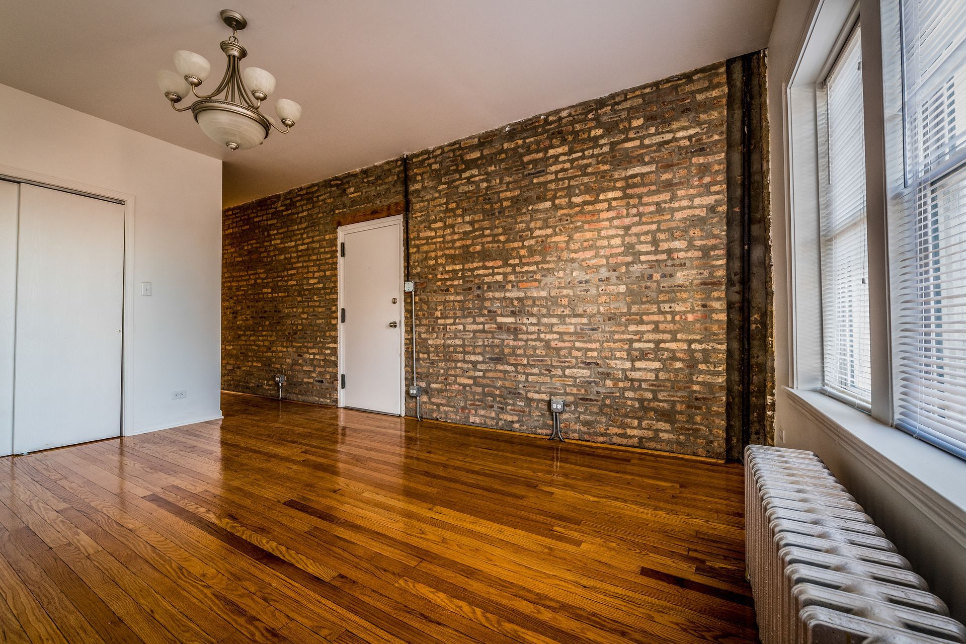 Empty room with hardwood floors, exposed brick wall, white door, and window with blinds.