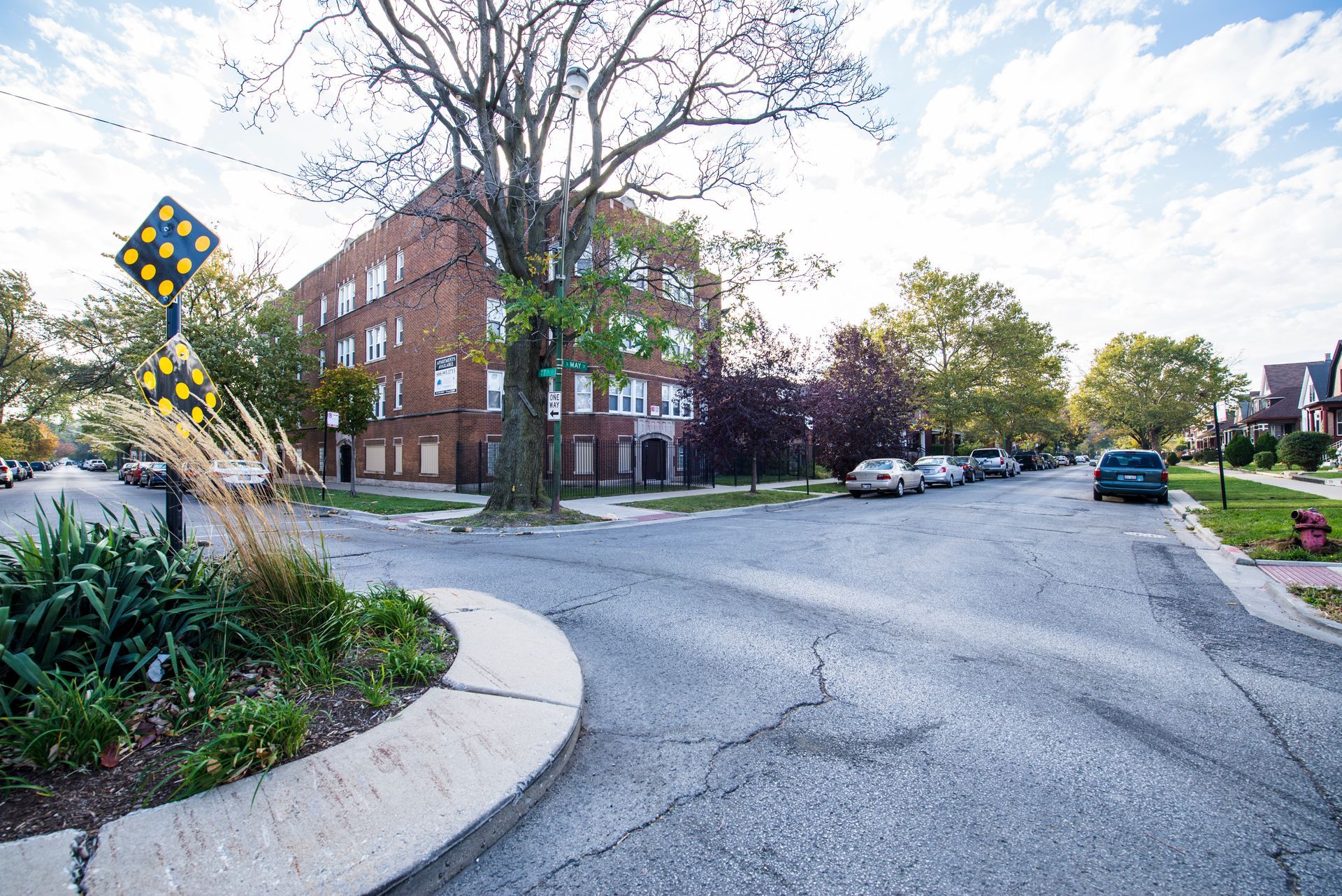 Street corner with a large brick building. A yield sign stands near greenery. Cars line the street.