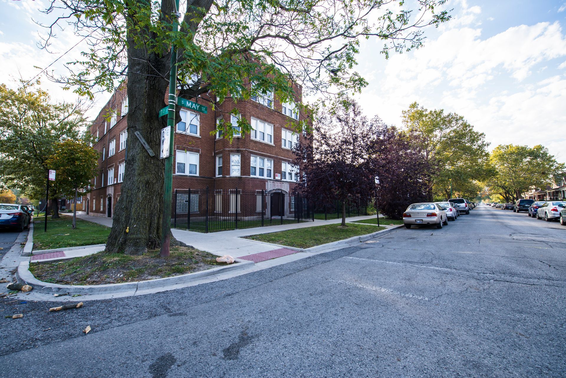 Brick apartment building on a street corner, cars parked along the street, trees, and blue sky.