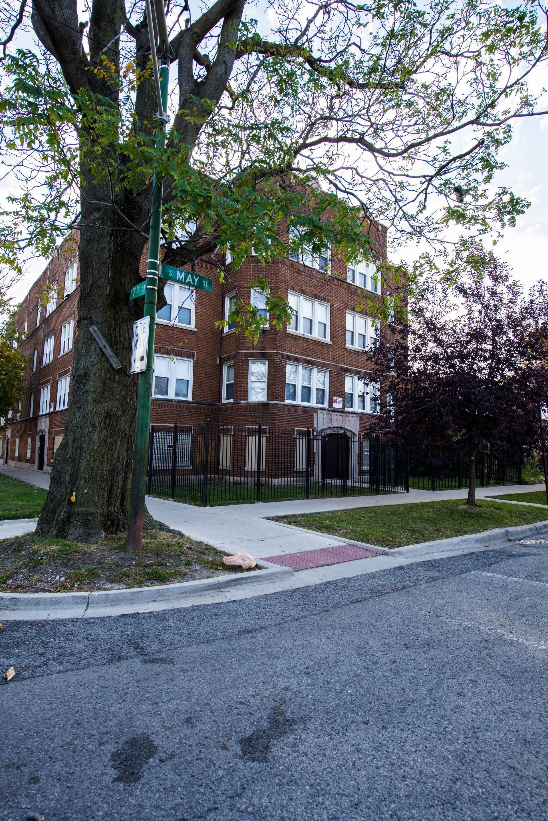 Brick apartment building on a corner lot, next to a large tree and sidewalk, overcast sky.