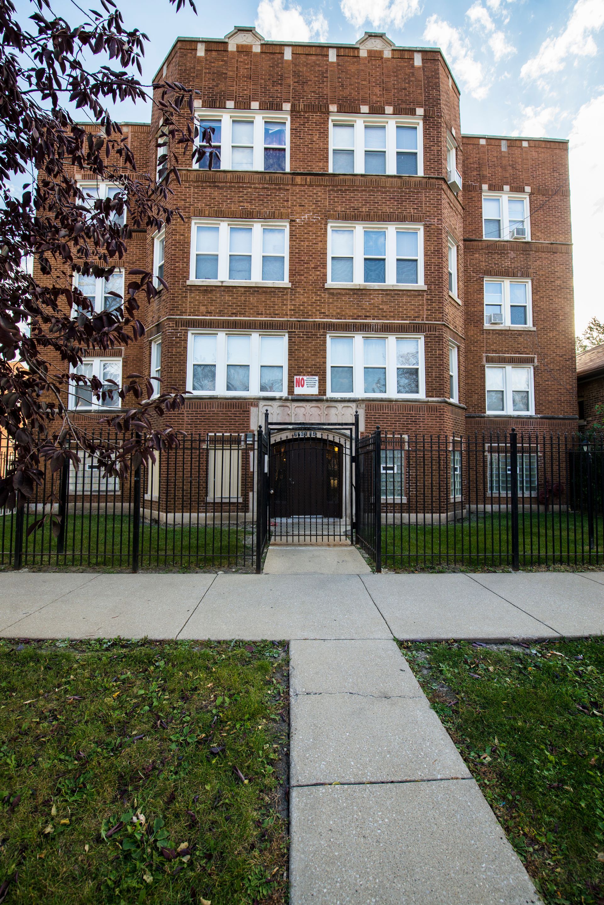 Red brick apartment building with white-framed windows, gated entrance, and a sidewalk.