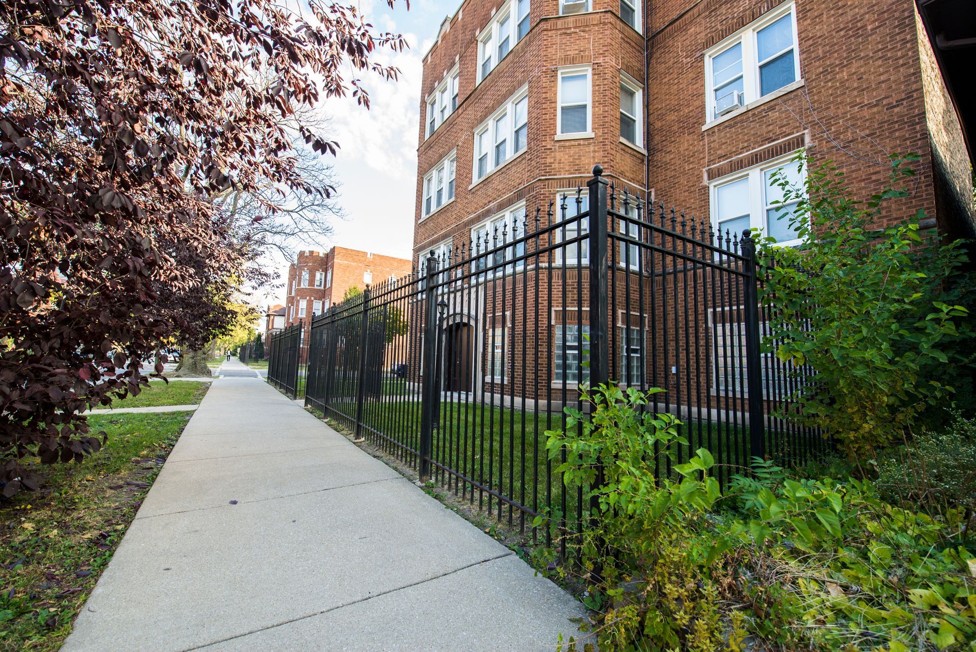 Sidewalk next to a black fence, with red brick apartment building. Lush greenery.