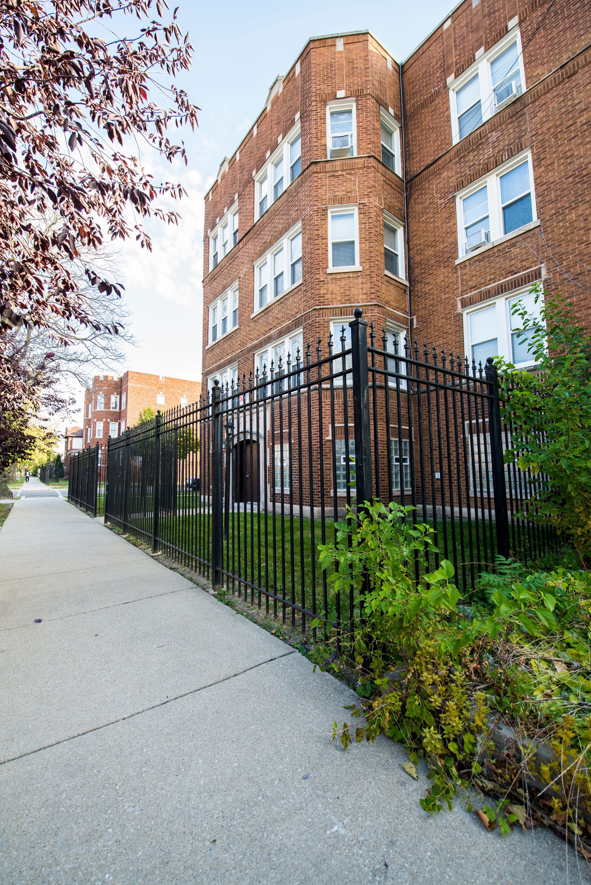 Brick apartment building with black fence along a sidewalk, surrounded by trees and greenery.