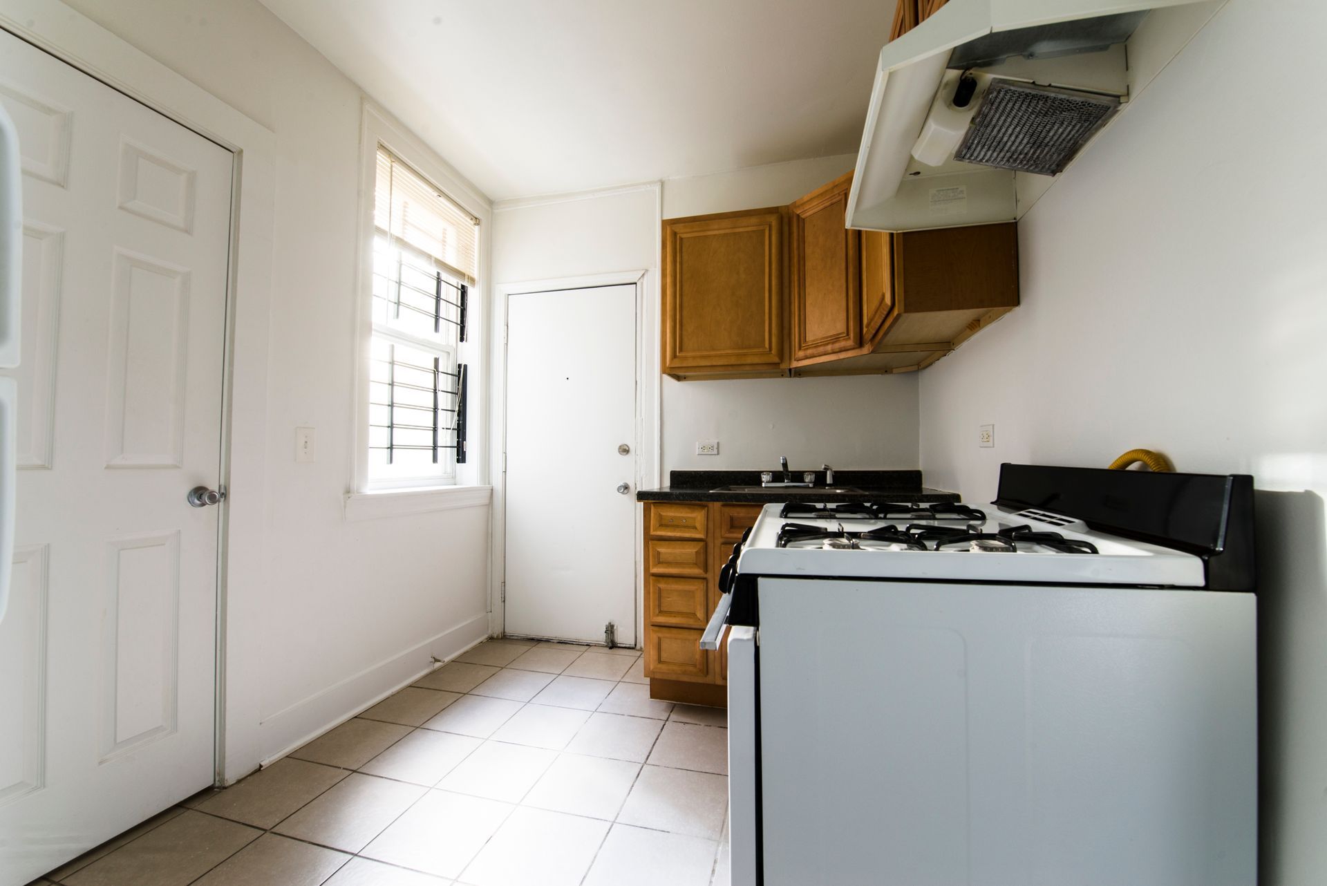 Small, white kitchen with wood cabinets, stove, sink, window, and two white doors.