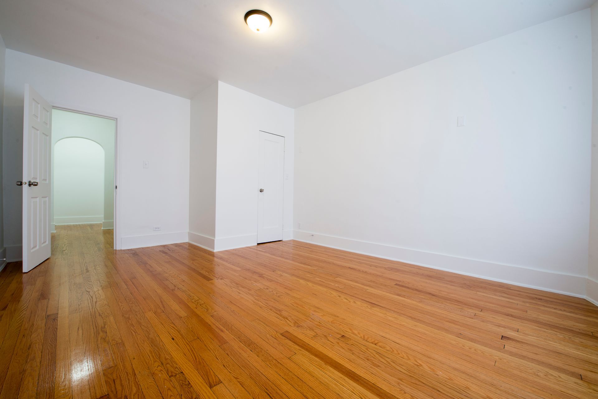 Empty room with hardwood floors, white walls, and an open doorway to another room.