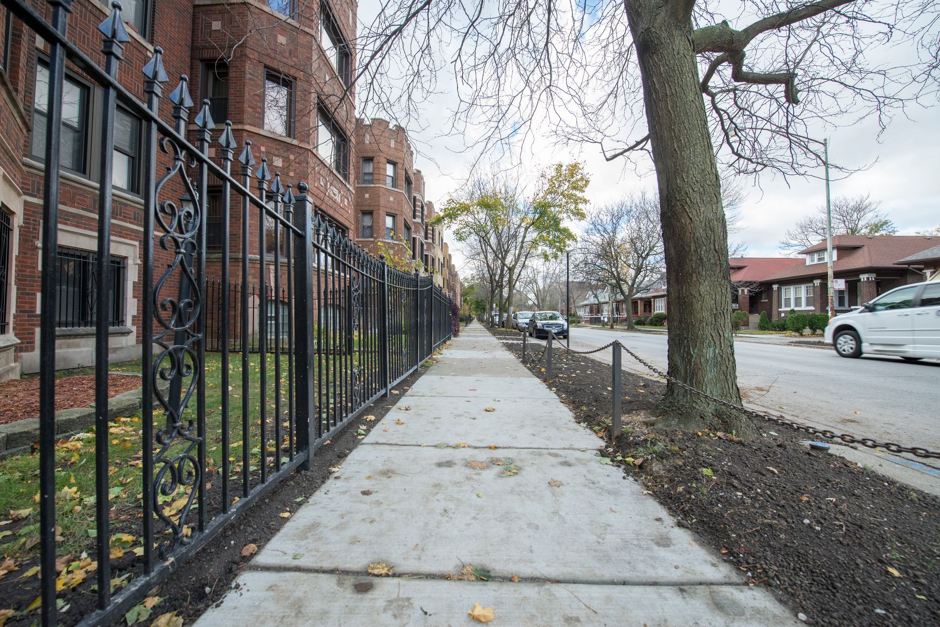 Sidewalk with black fence, brown brick buildings, and parked cars on a city street.