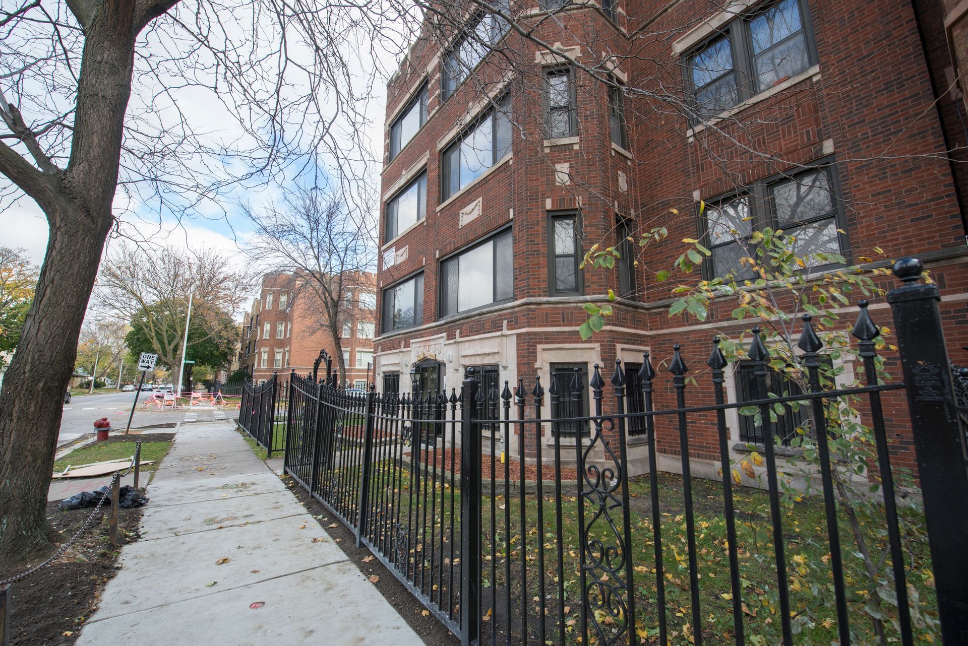 Brick apartment building with black wrought-iron fence along a sidewalk, trees, and street.