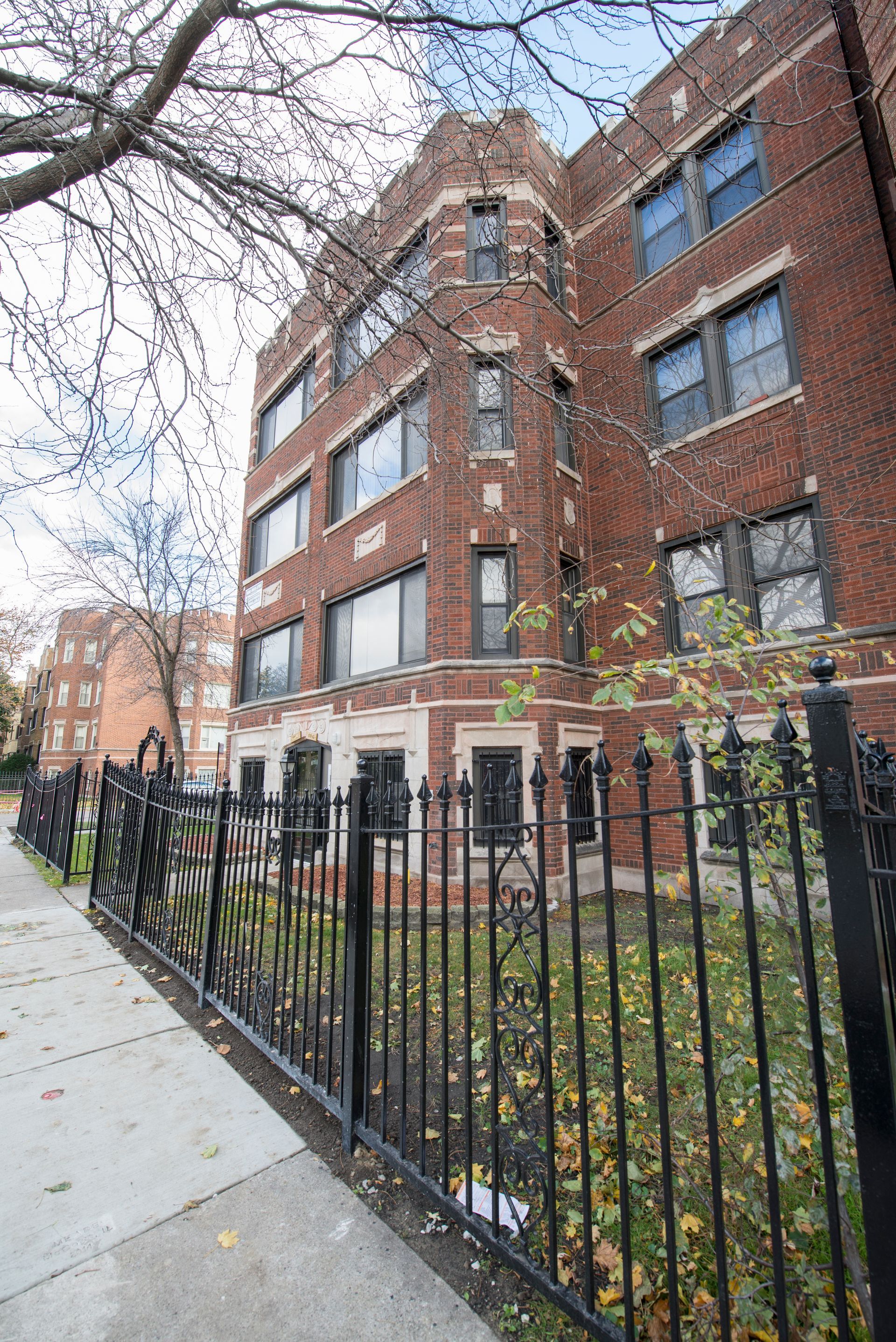 Brick apartment building with black iron fence, sidewalk, and trees.