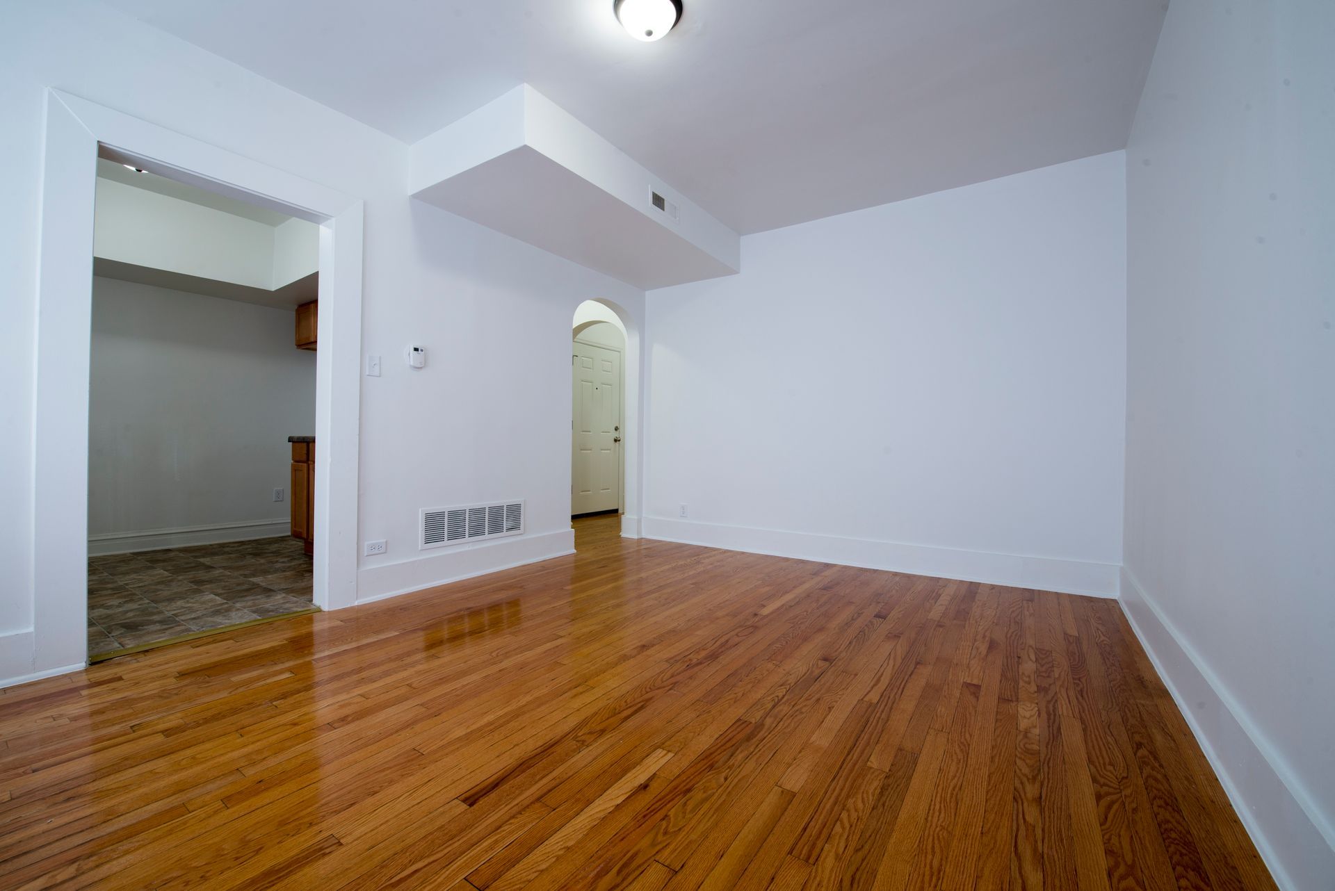 Empty room with hardwood floors, white walls, and a doorway leading to a kitchen.