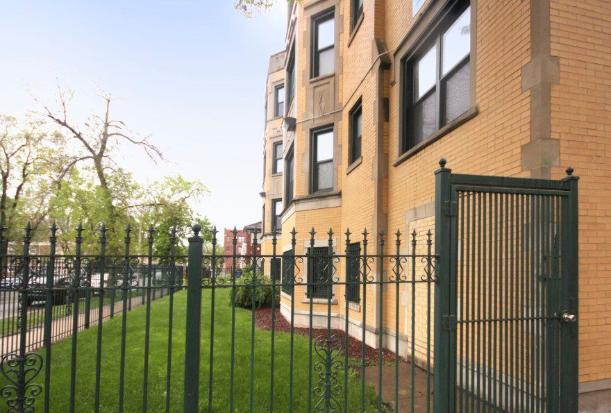 Green iron fence and gate in front of a tan brick building with dark window frames.