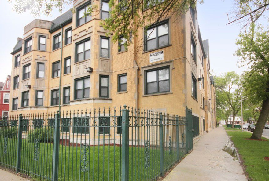 A three-story brick apartment building with a wrought iron fence and sidewalk.