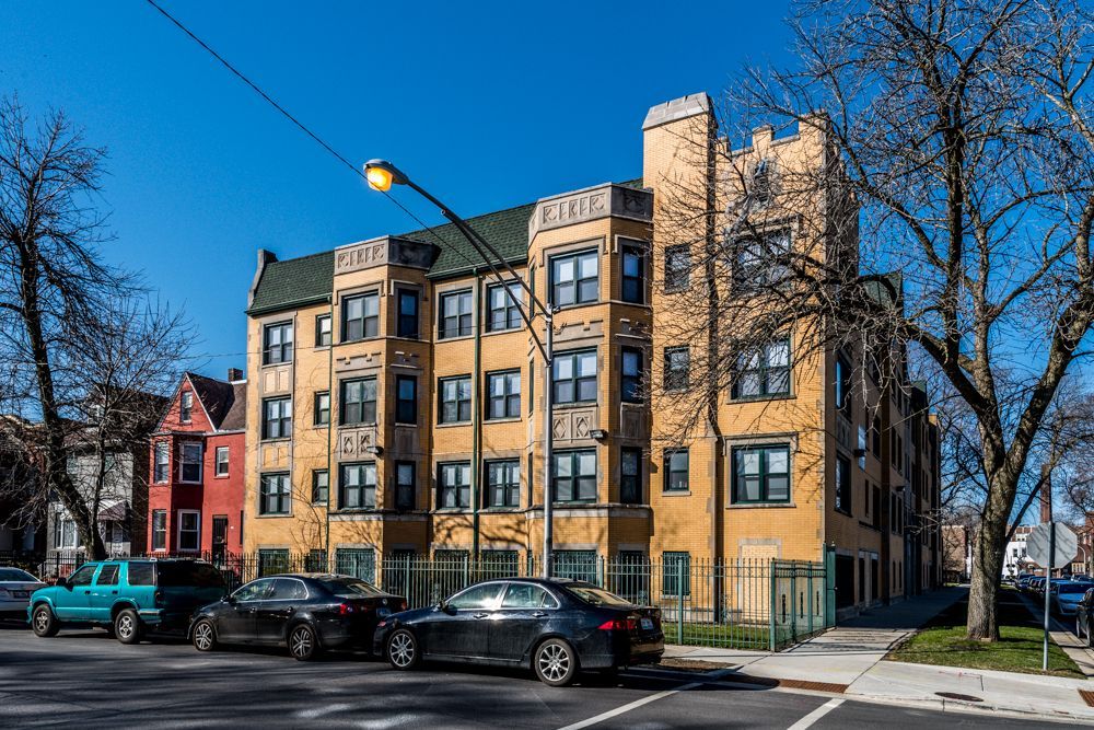 Yellow multi-story apartment building with cars parked in front. Blue sky.