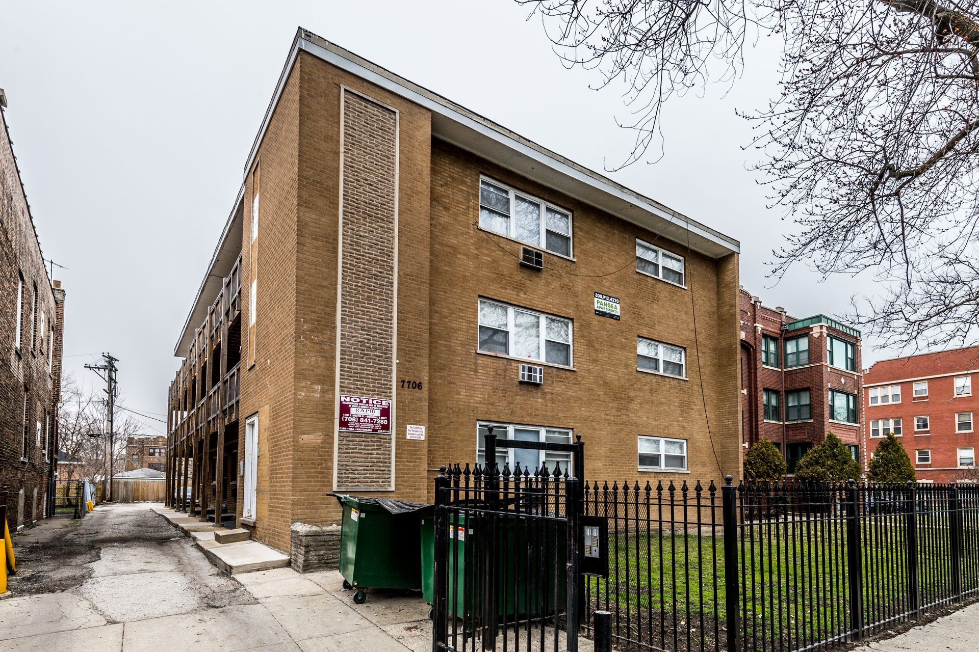 Three-story brick apartment building with dark fence and trash cans in front.