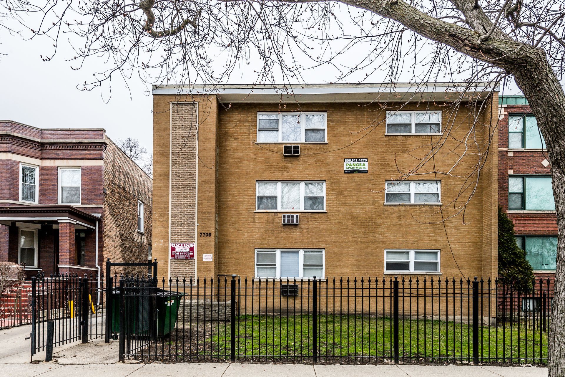 Three-story brick apartment building behind a black metal fence, on a cloudy day.