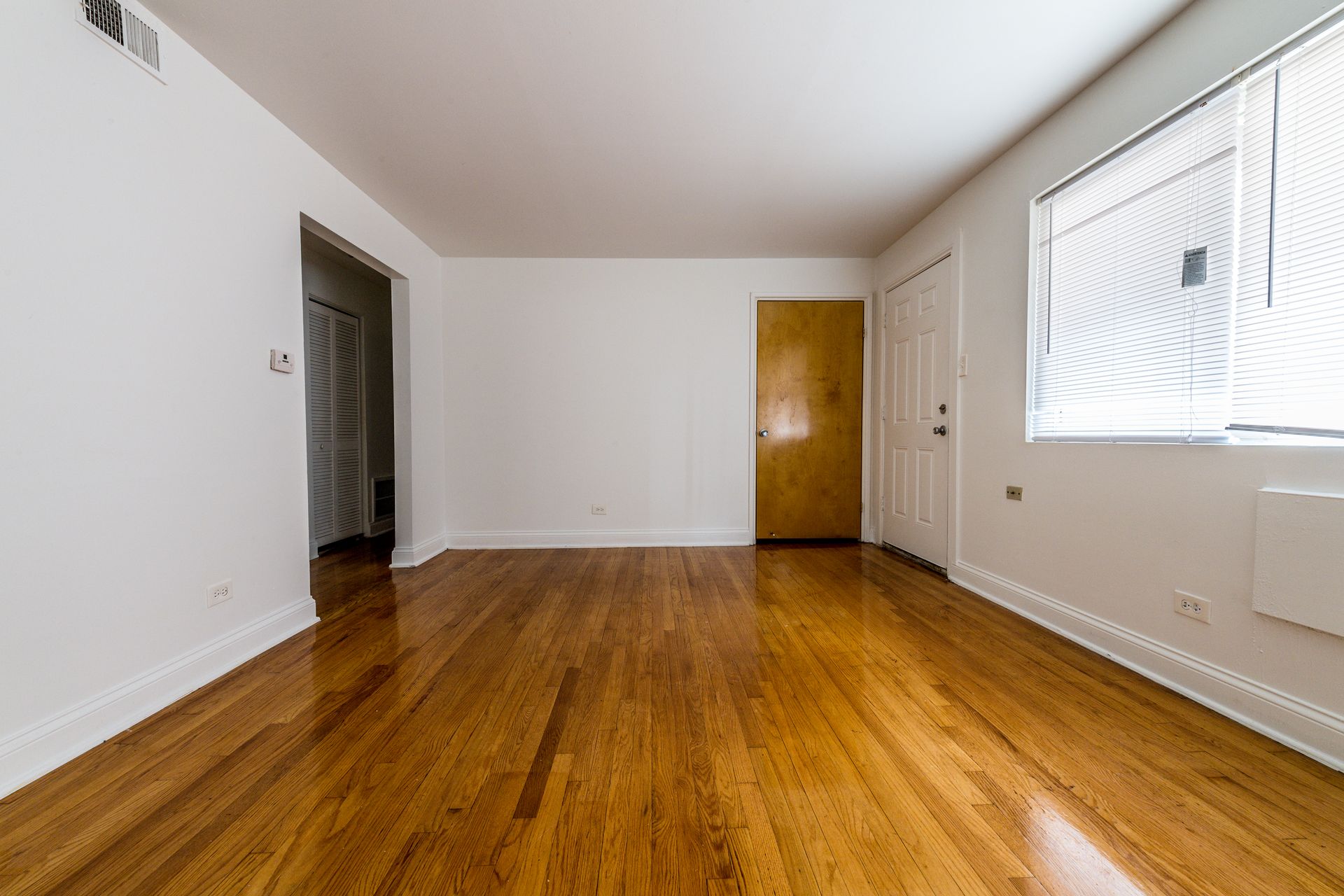 Empty room with hardwood floors, white walls, and a window with blinds.