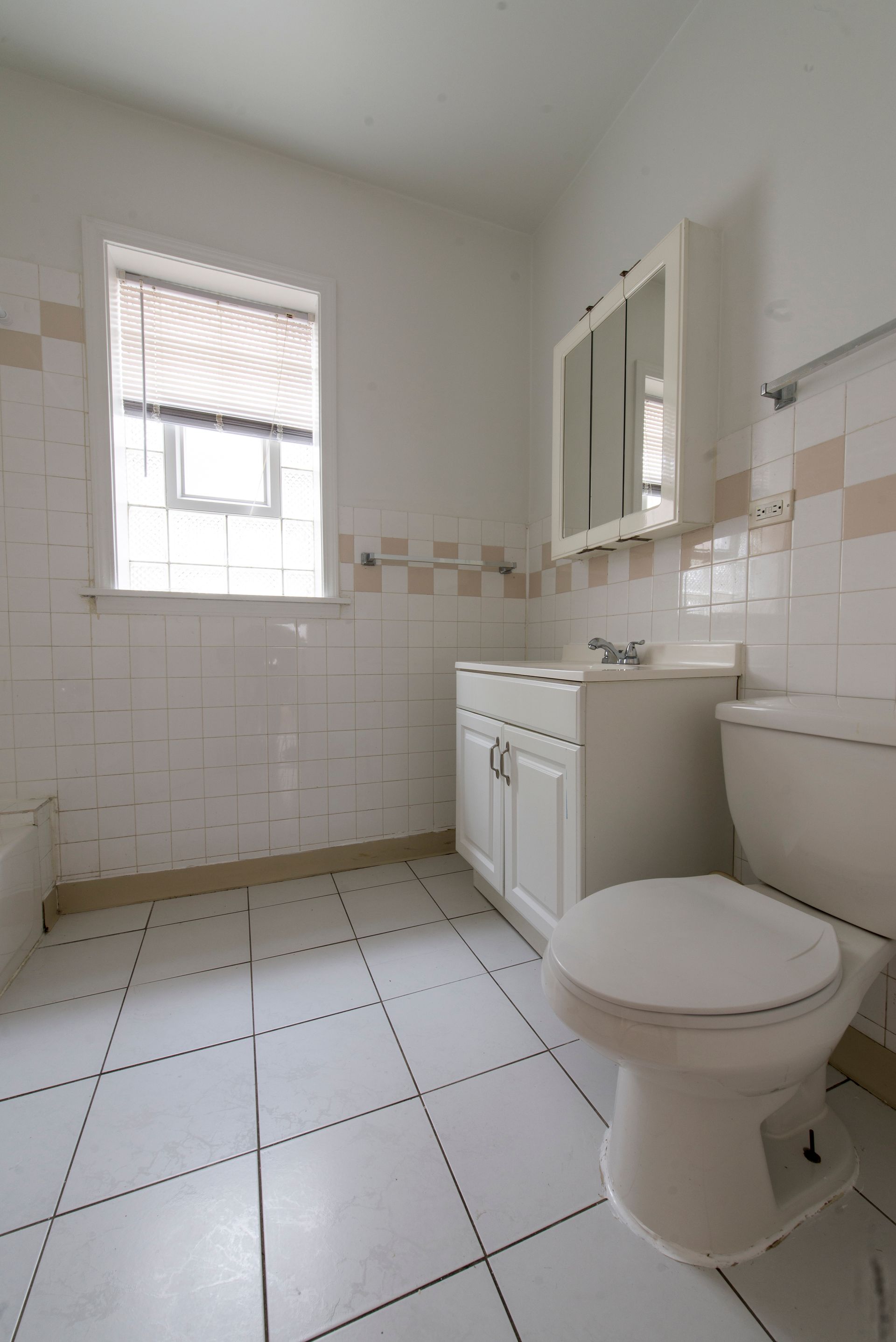 Small, white-tiled bathroom with toilet, sink, and window with blinds.