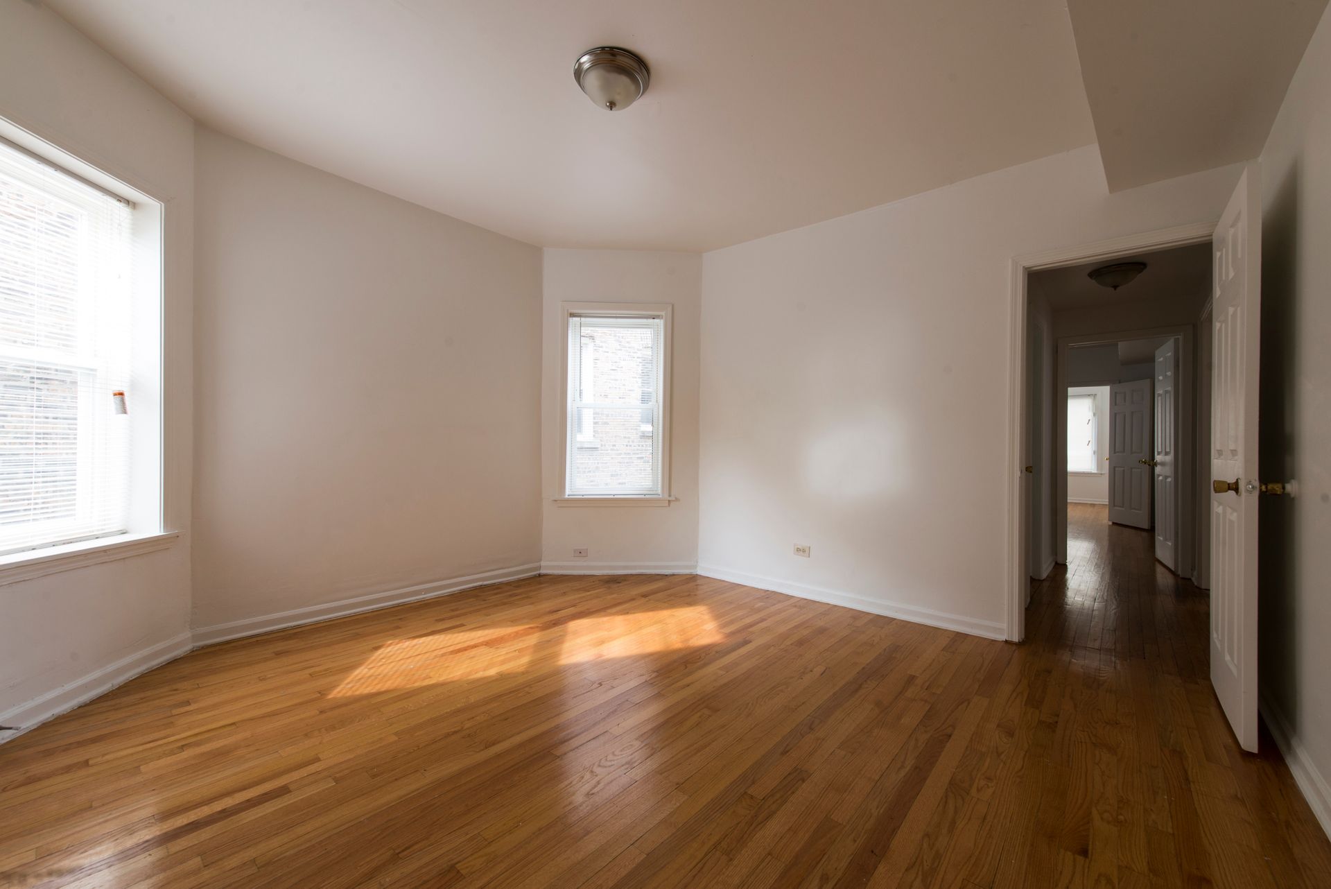 Empty room with hardwood floors, white walls, and sunlight streaming through windows.