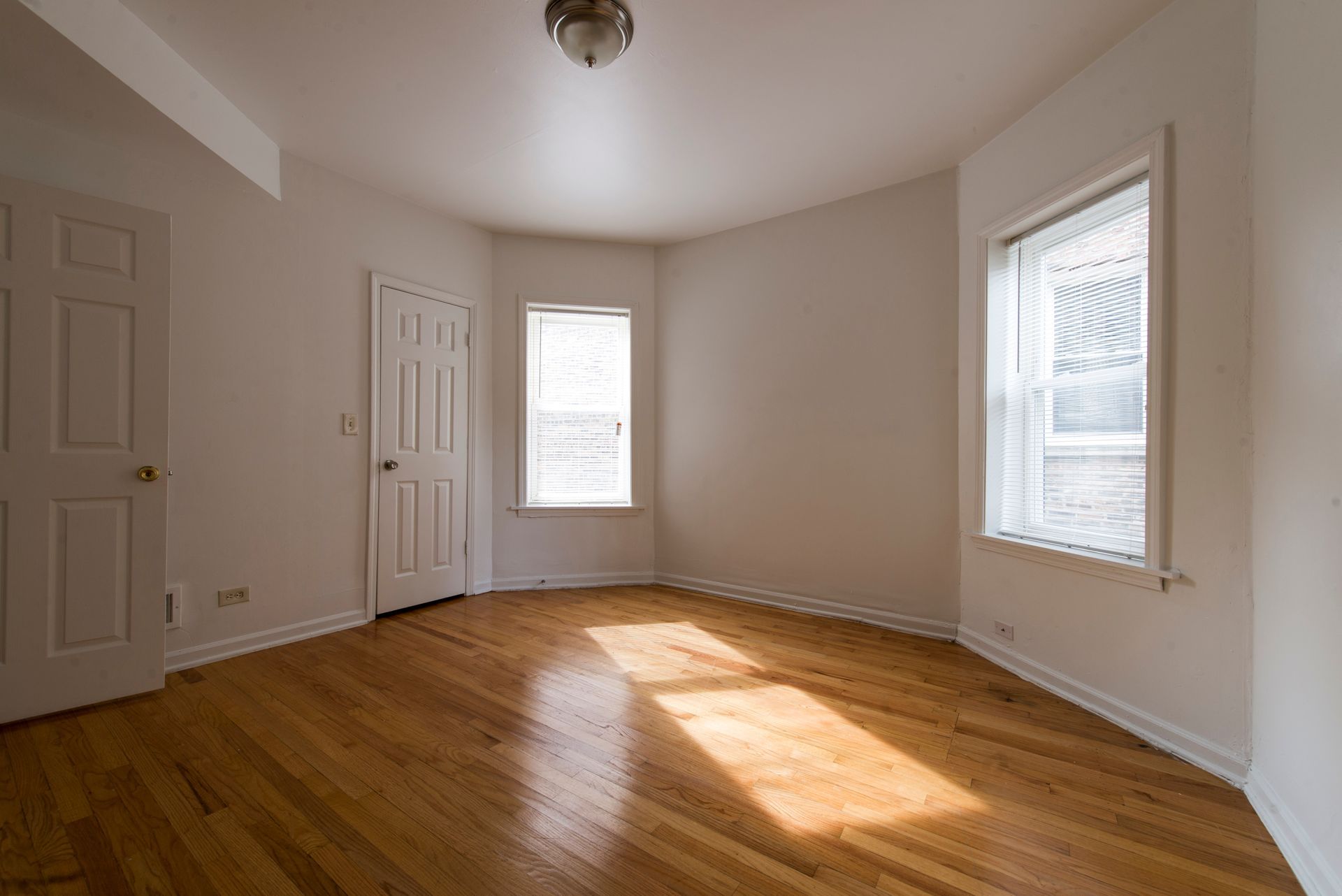 Empty room with hardwood floors, two windows, and a closed door. Sunlight streams through the windows.