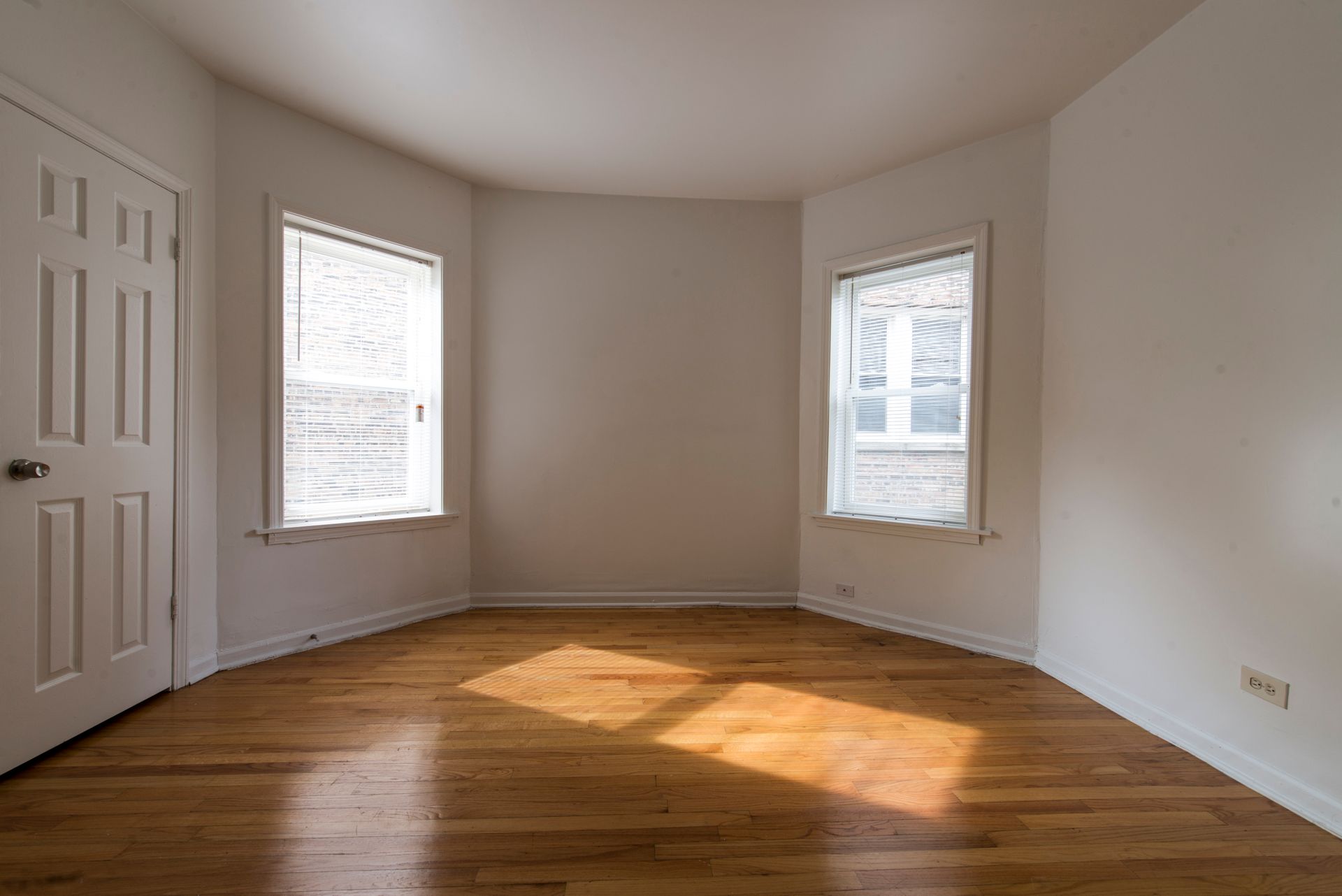 Empty room with hardwood floors, white walls, two windows, and a closed door.