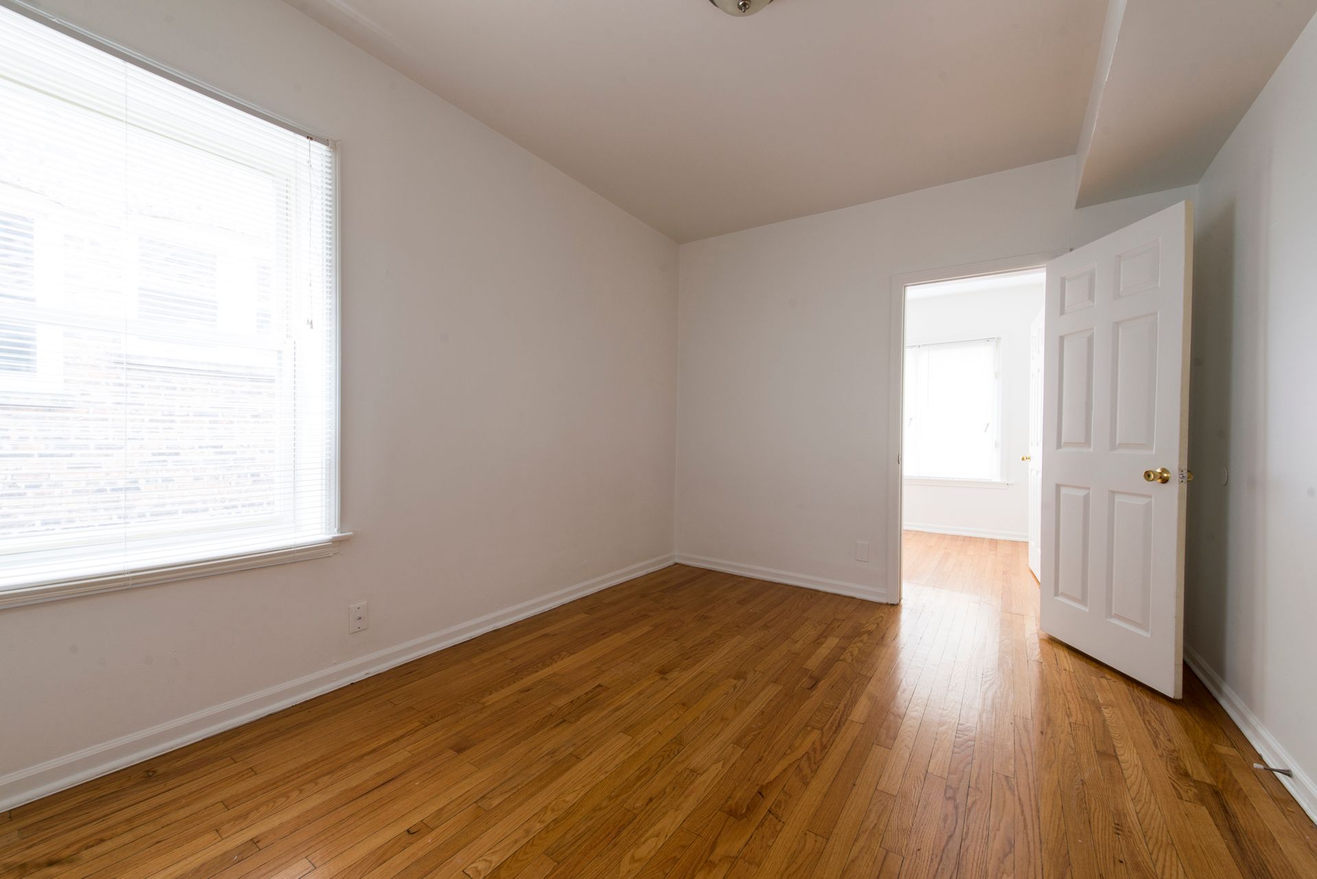 Empty room with wood floor, white walls, window with blinds, and open door.