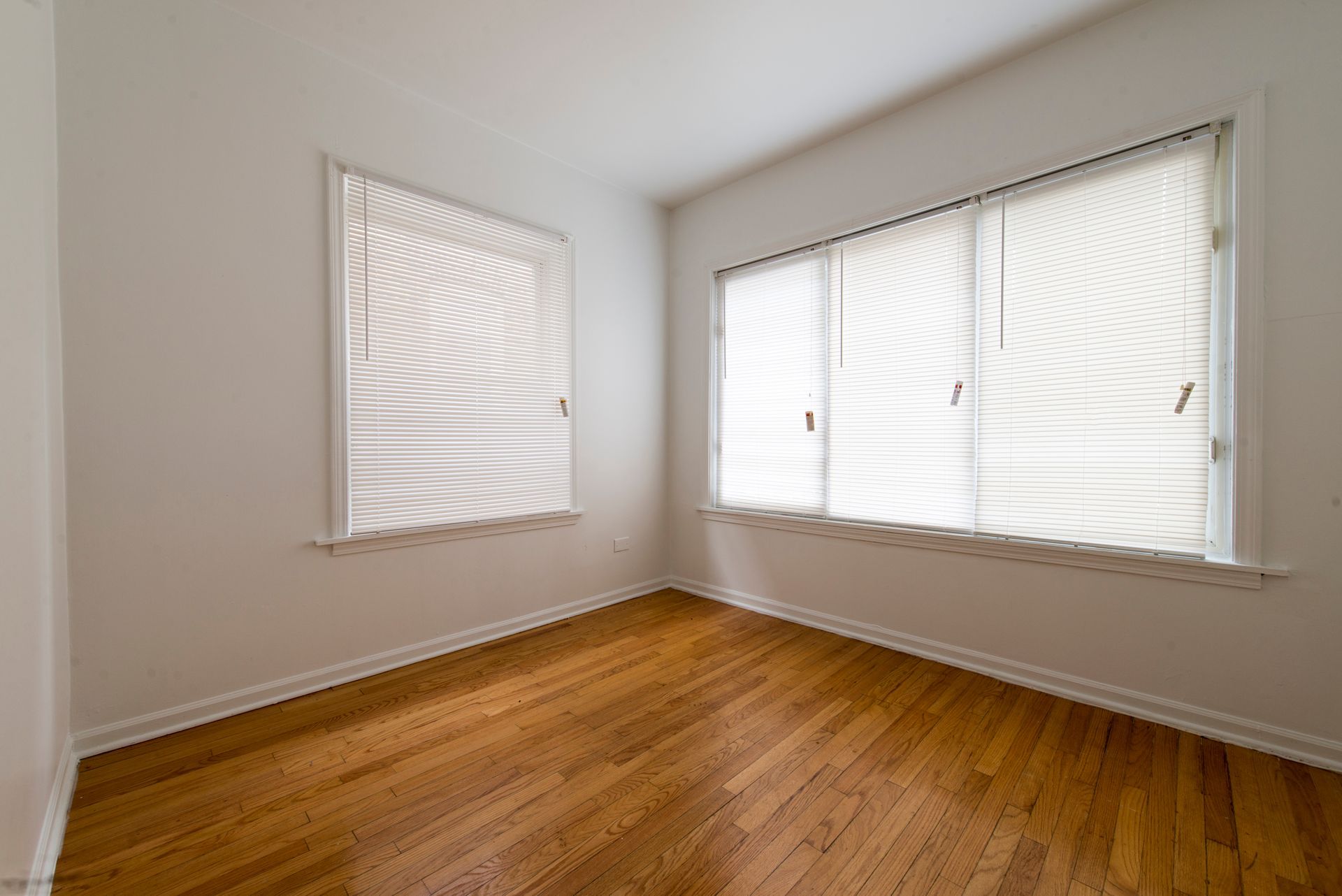 Empty room with hardwood floors, two windows with white blinds, and white walls.
