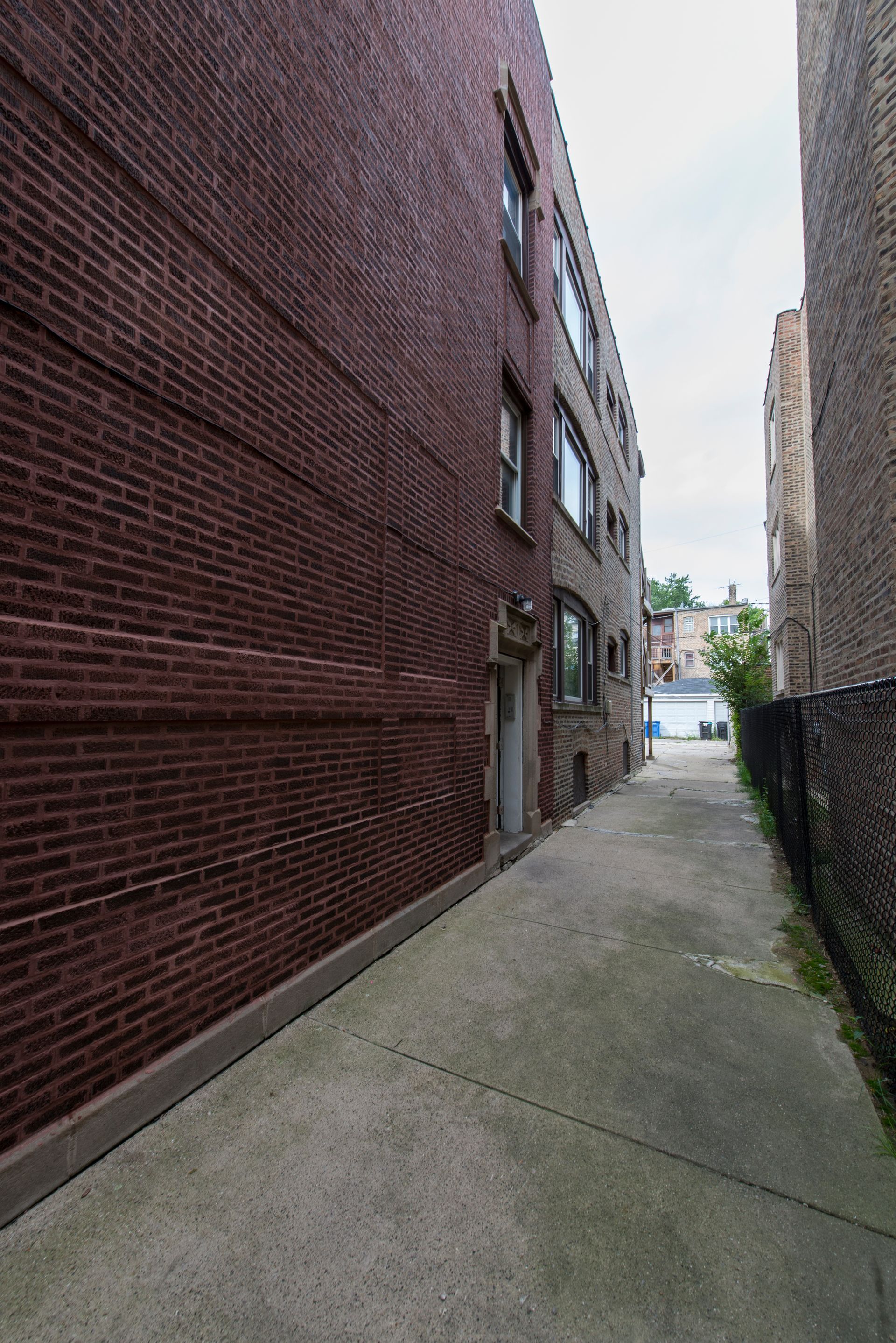 Alleyway between brick buildings, paved with concrete. A metal fence and foliage on the right.