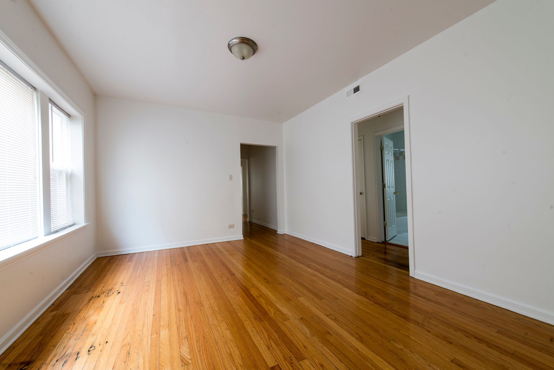 Empty room with hardwood floors, white walls, and doorway. Window with blinds on the left.
