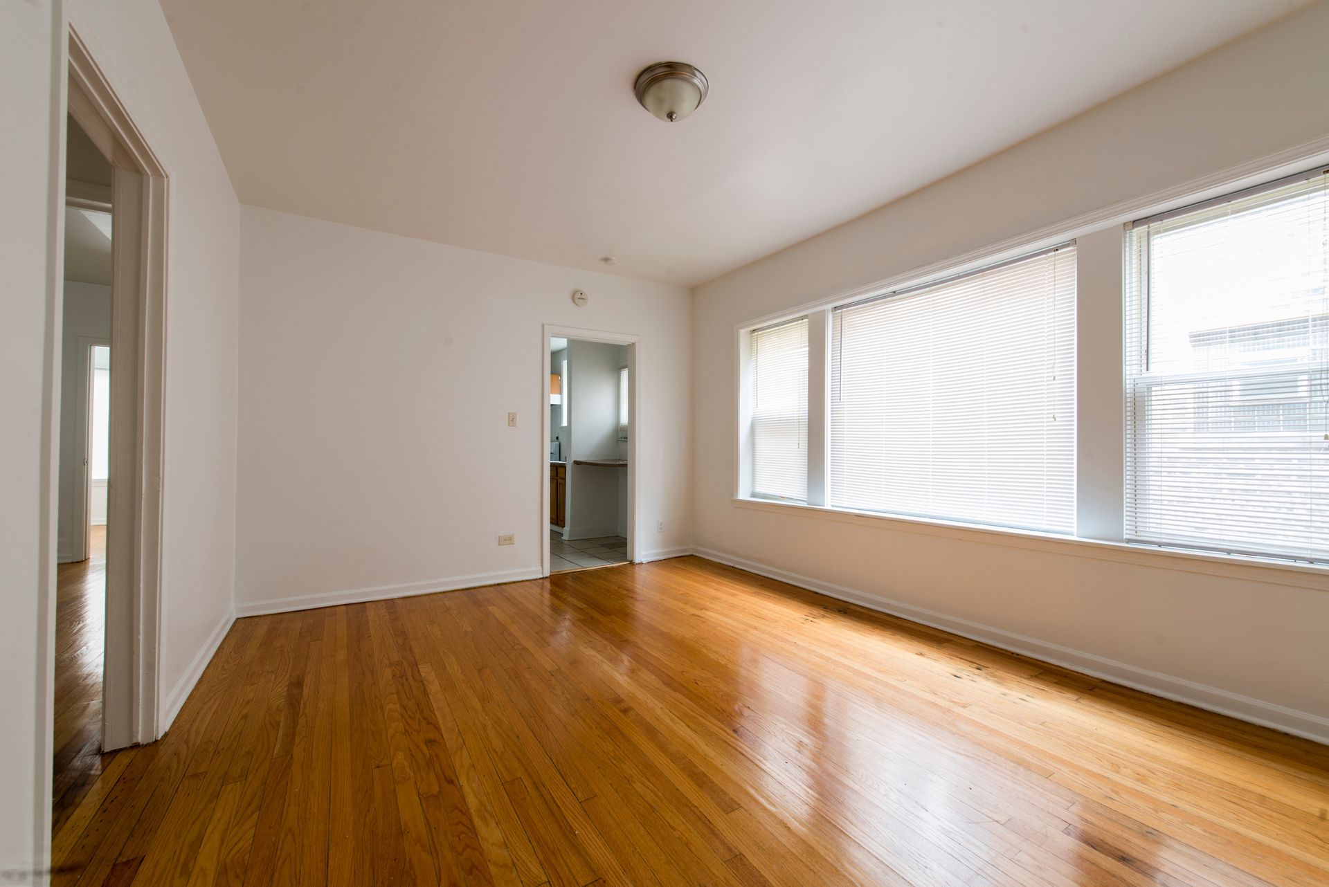 Empty room with wood floors, white walls, windows with blinds, and a doorway.