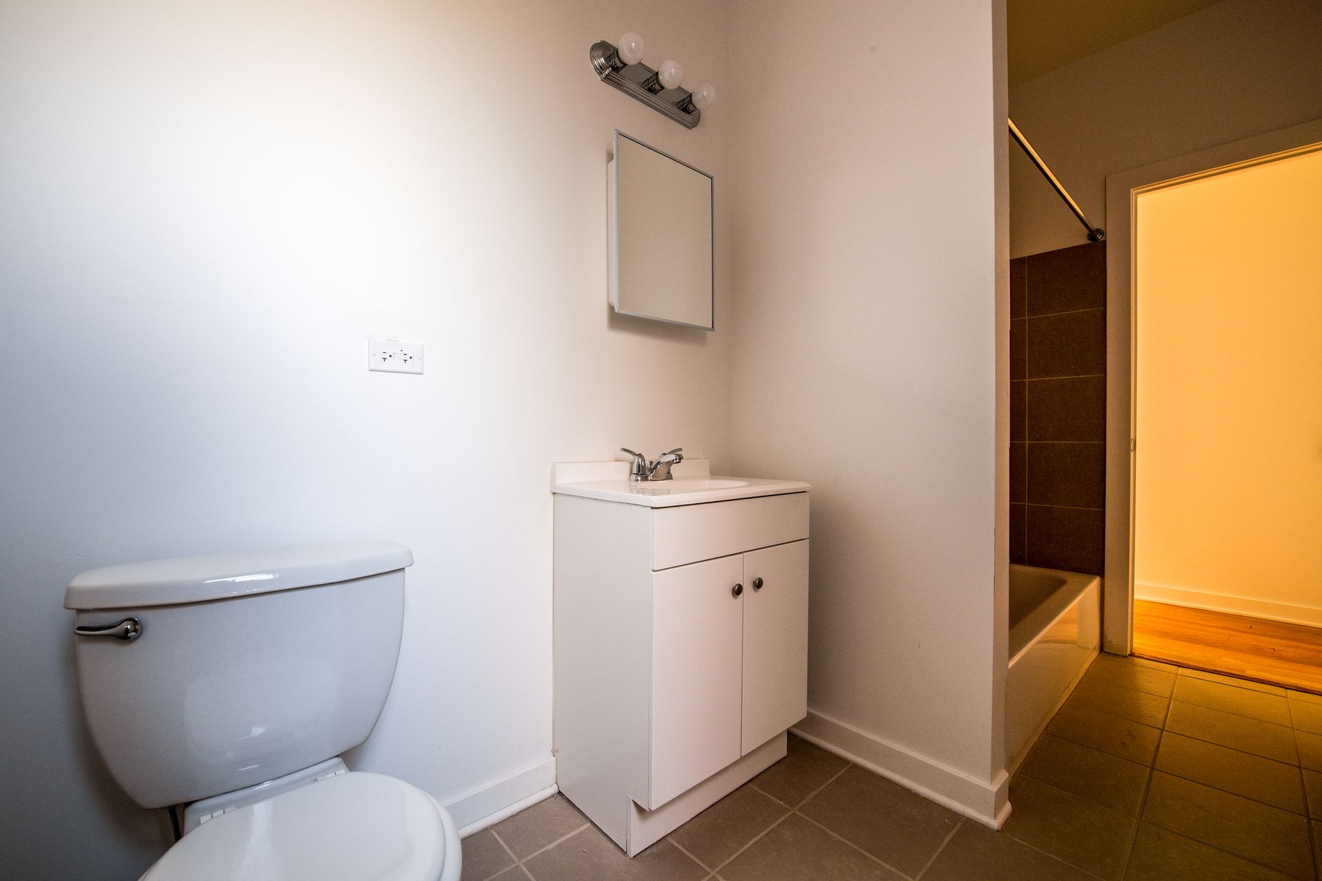 Bathroom with toilet, vanity, and open doorway to a bathtub. White walls and neutral floor.