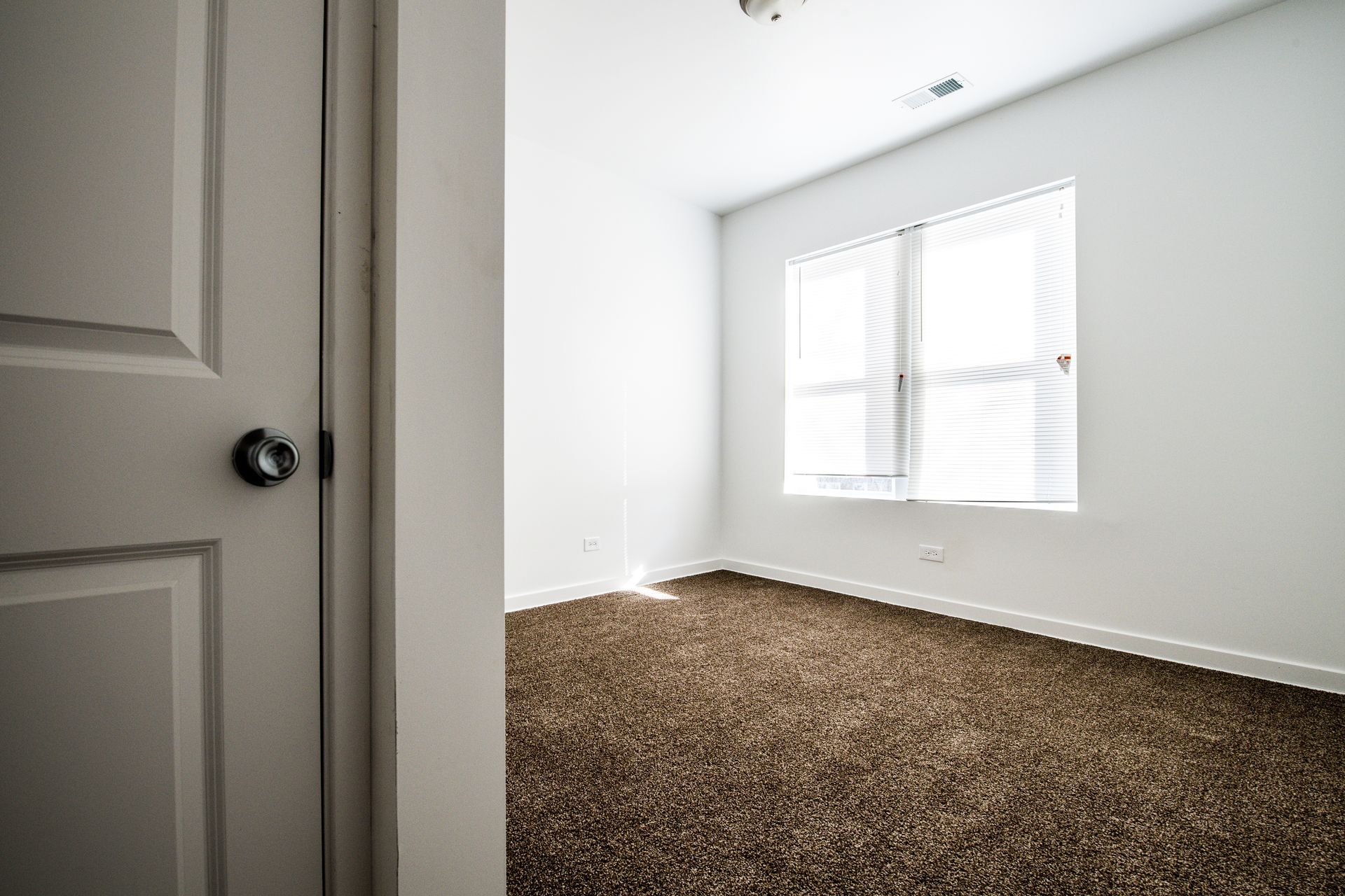 Interior view of a mostly empty room with brown carpet, white walls, and a window with blinds. A door is partially visible.