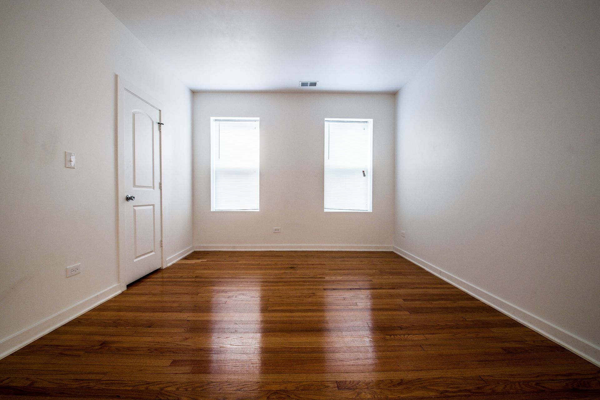 Empty room with hardwood floors, white walls, two windows, and a closed door.