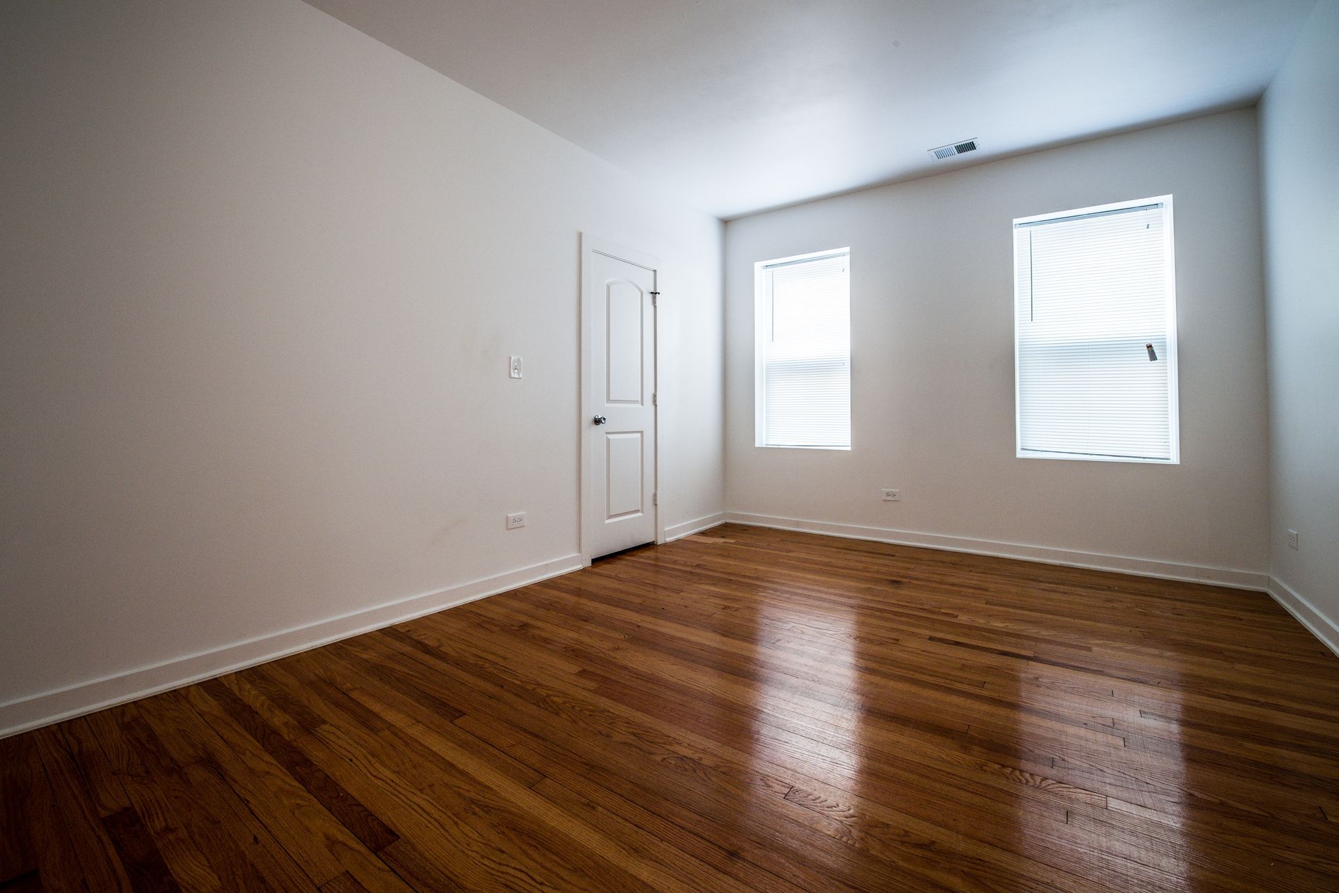 Empty room with hardwood floors, white walls, two windows with blinds, and a closed door.