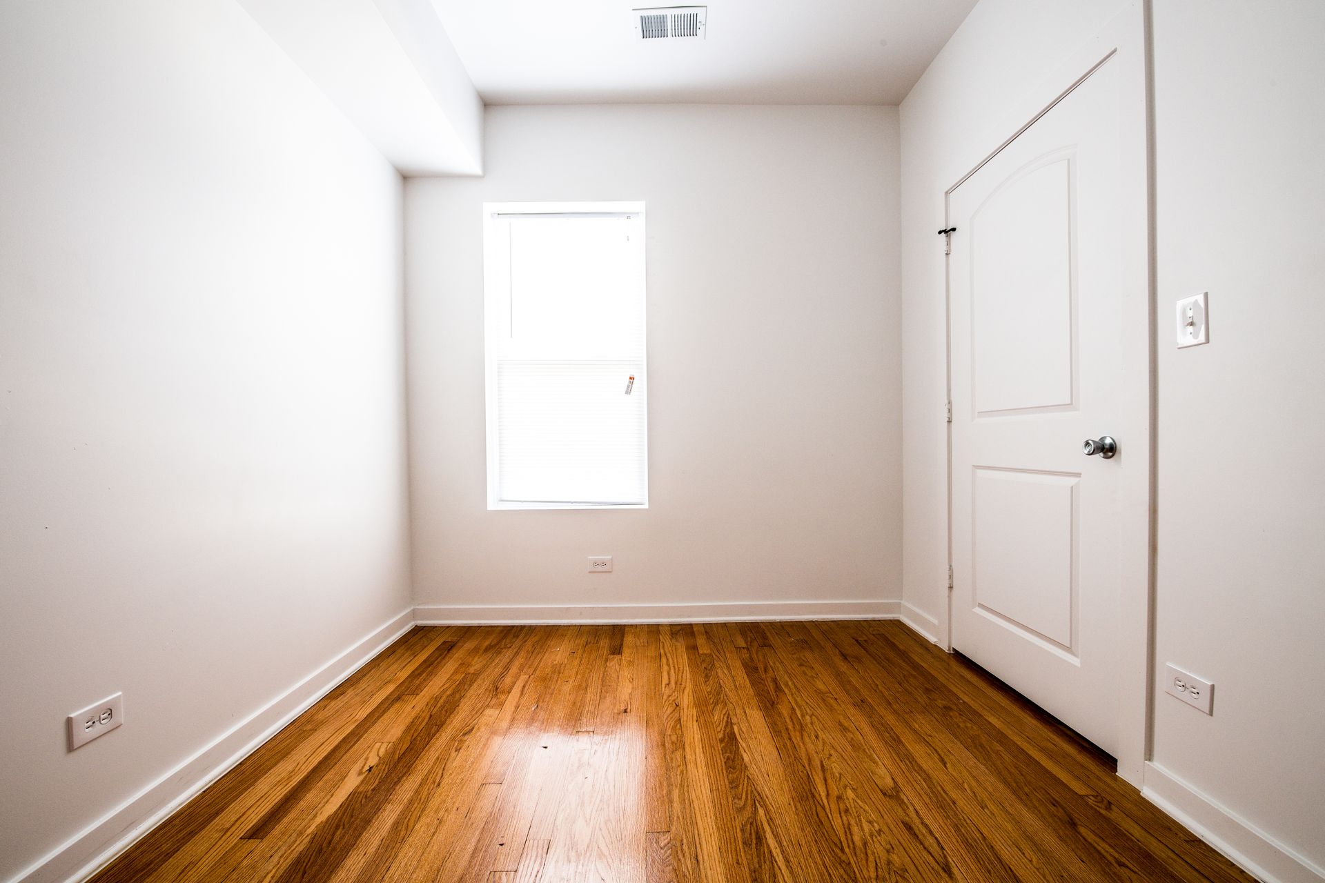 Empty room with wood floors, white walls, two windows with blinds, and a closed door.