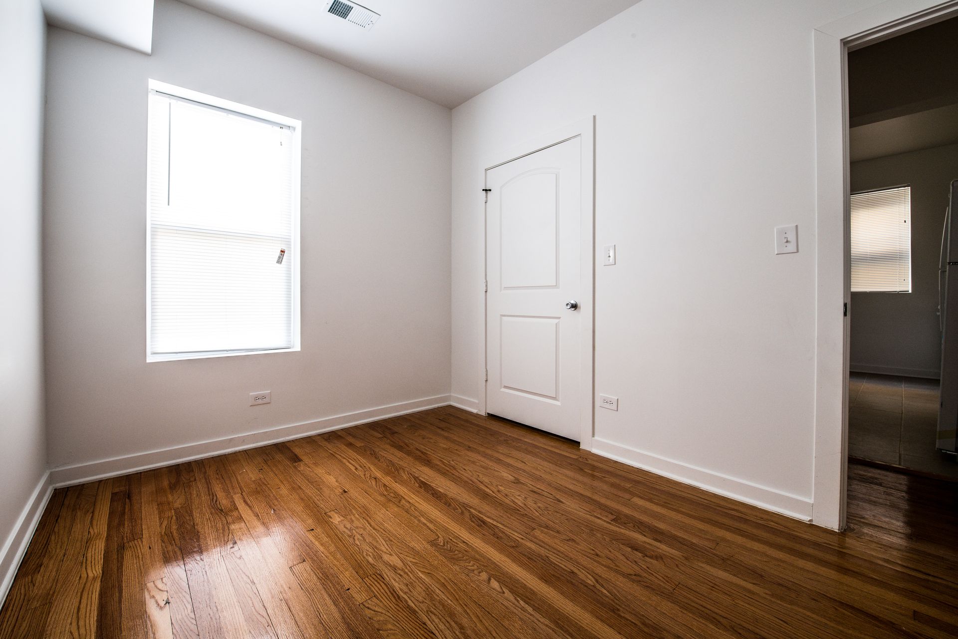 Empty room with hardwood floors, a window with blinds, and a closed door.