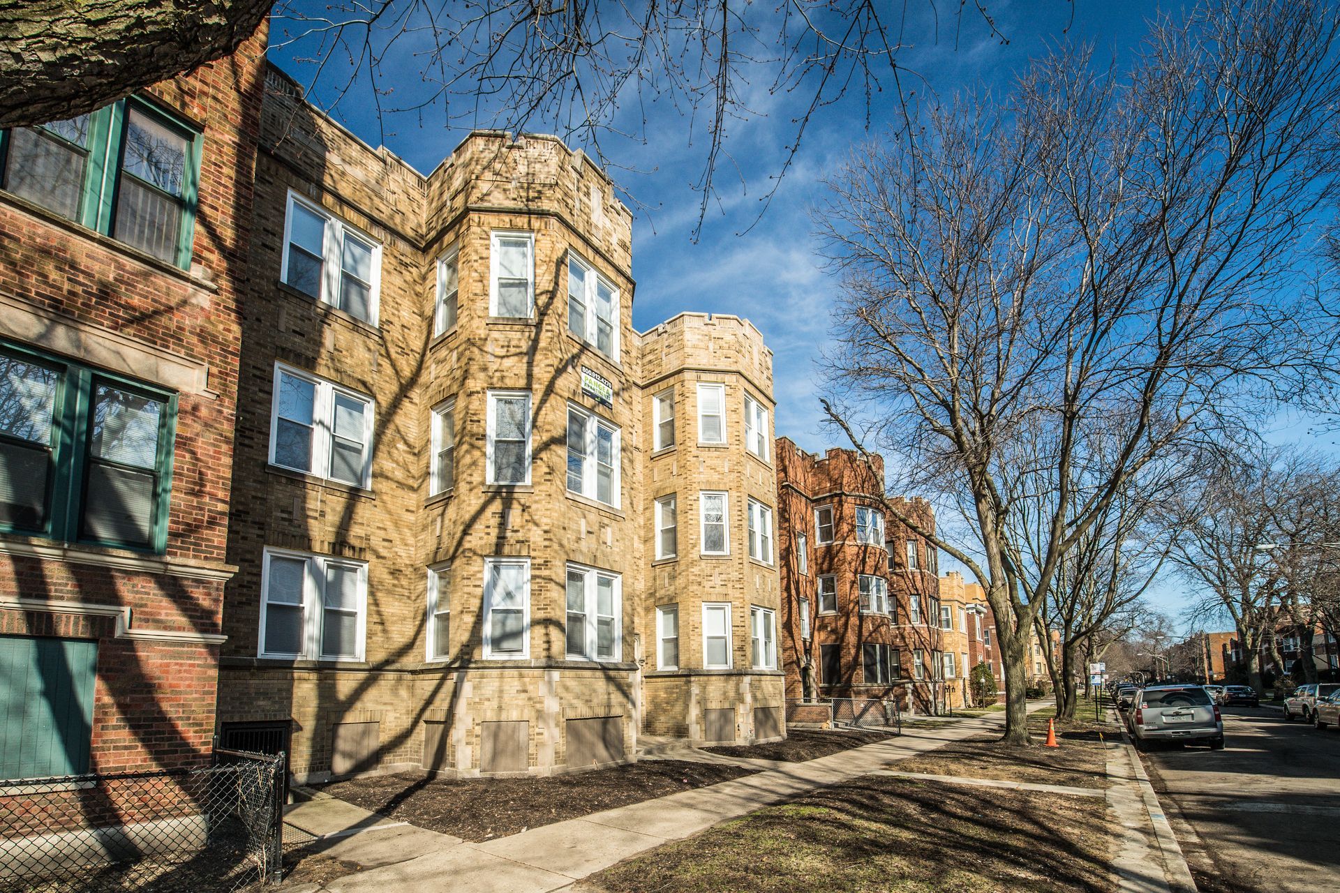 Row of brick apartment buildings on a sunny street, shadows of trees.
