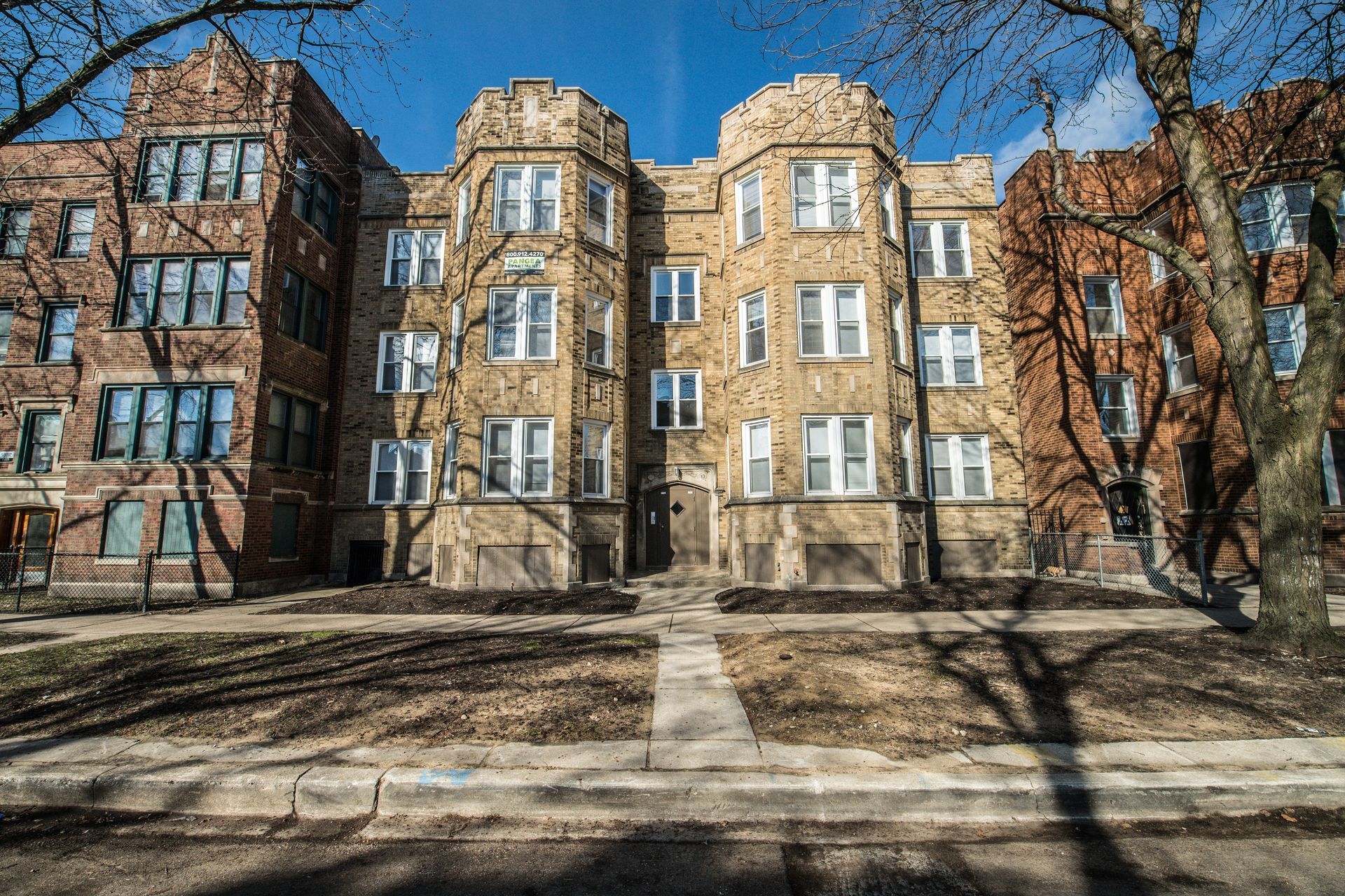 Multi-story brick apartment building on a sunny street, with trees in front.