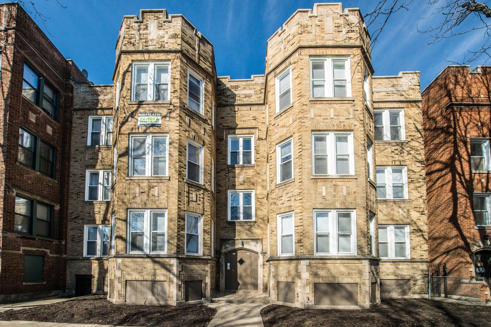 Three-story tan brick apartment building with architectural details; sunny day, blue sky.