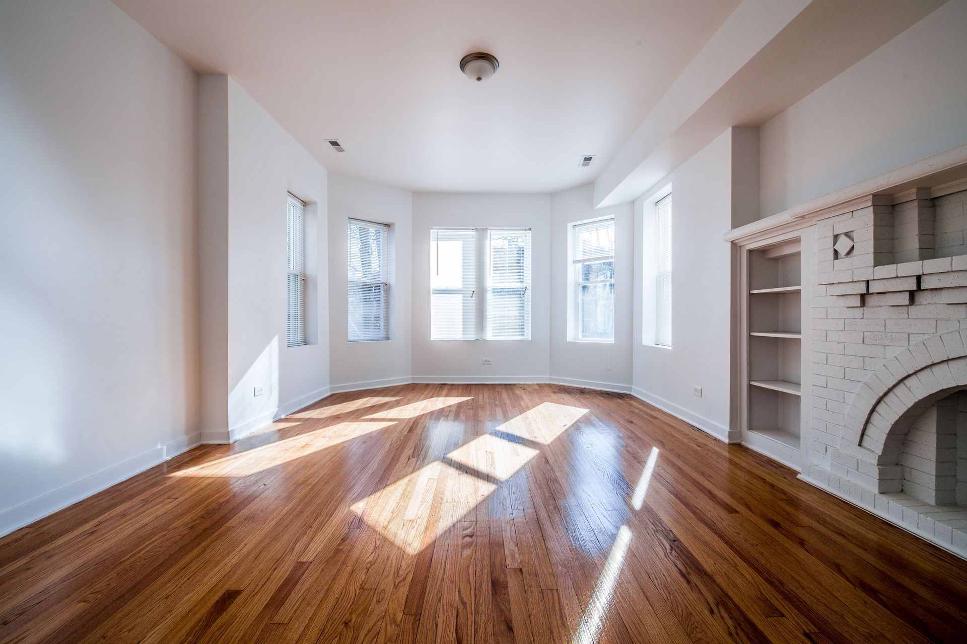 Empty room with hardwood floors, bay windows, and a fireplace. Bright sunlight streams in.
