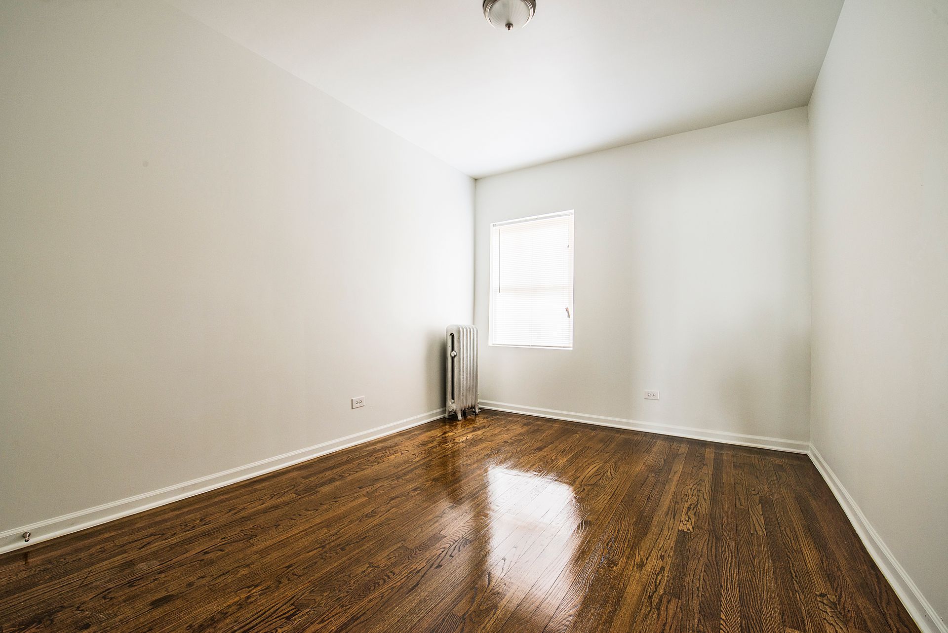 Empty room with hardwood floors, white walls, small window, and radiator.