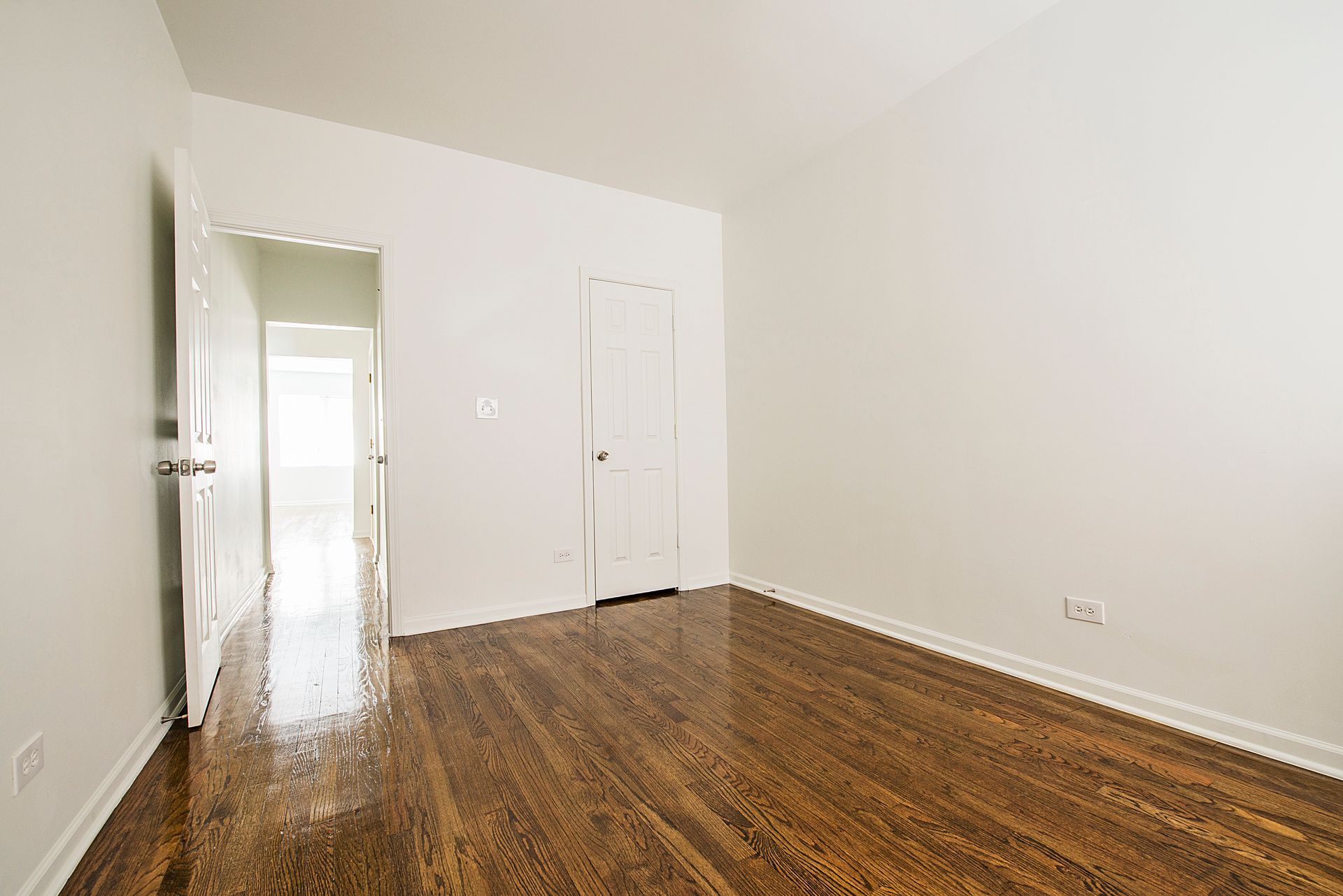 Empty room with hardwood floors, white walls, and a closed door. Another doorway leads to another room.