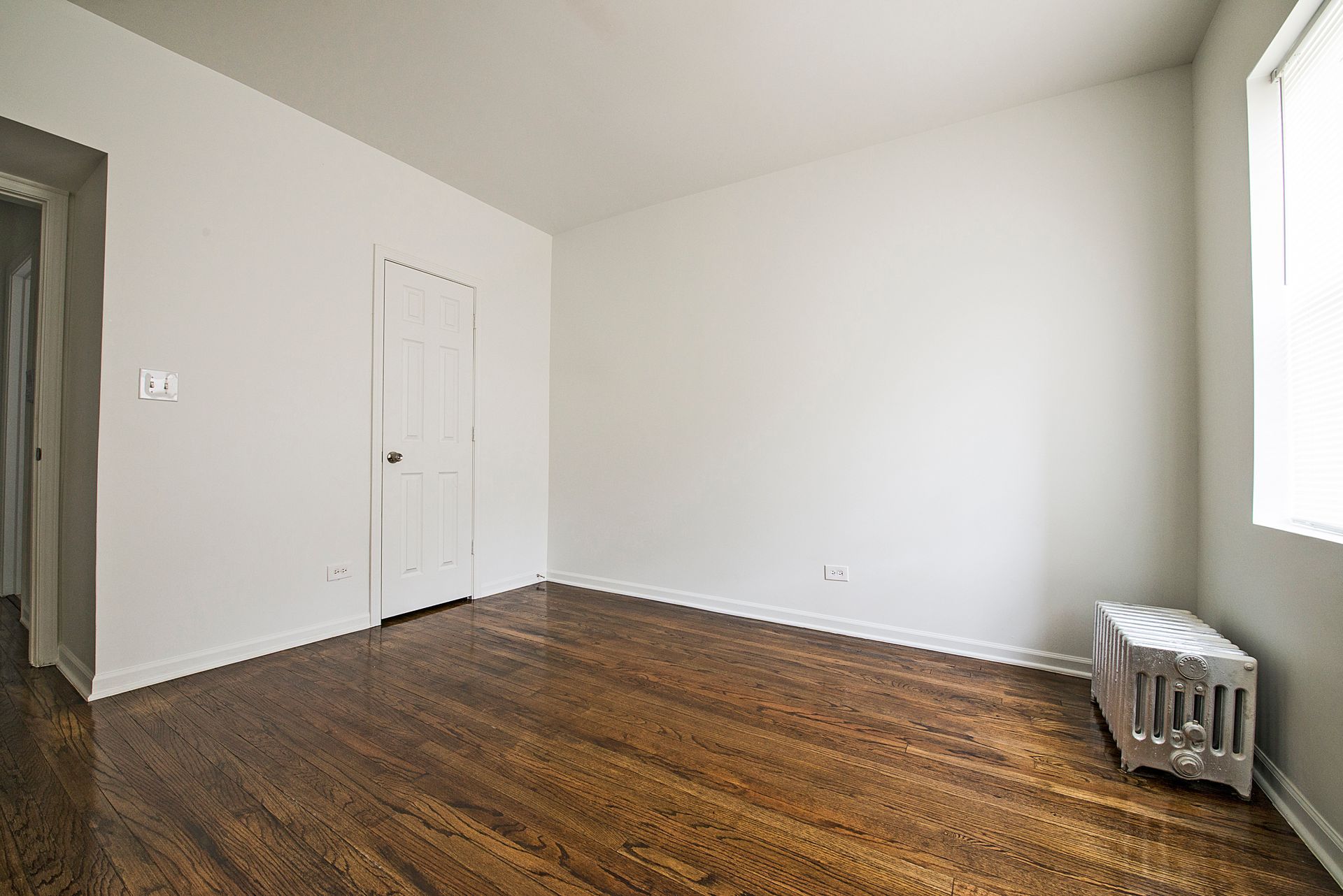 Empty room with hardwood floors, white walls, small door, window, and radiator.