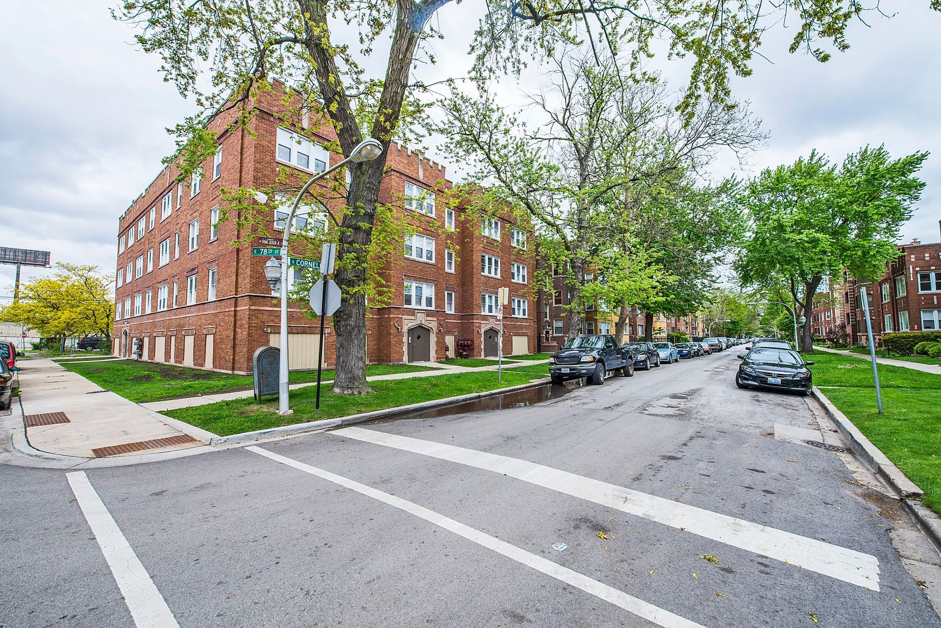 Brick apartment building on a street with parked cars and crosswalk lines, overcast sky.