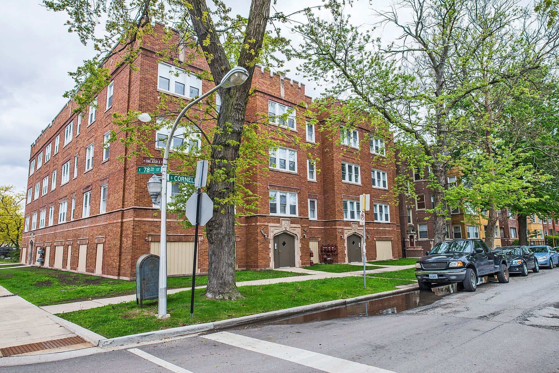 Brick apartment building on a street with parked cars and a large tree in front.