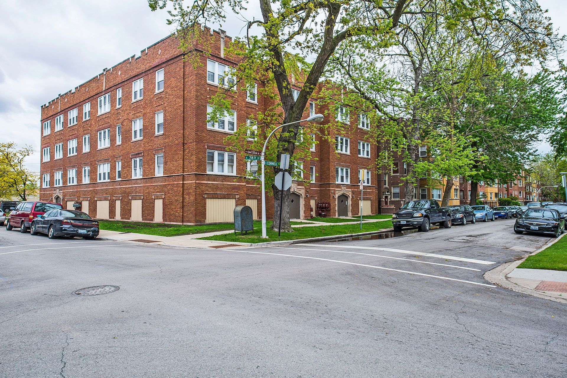 Brick apartment building at a street corner with parked cars and a tree. Overcast sky.