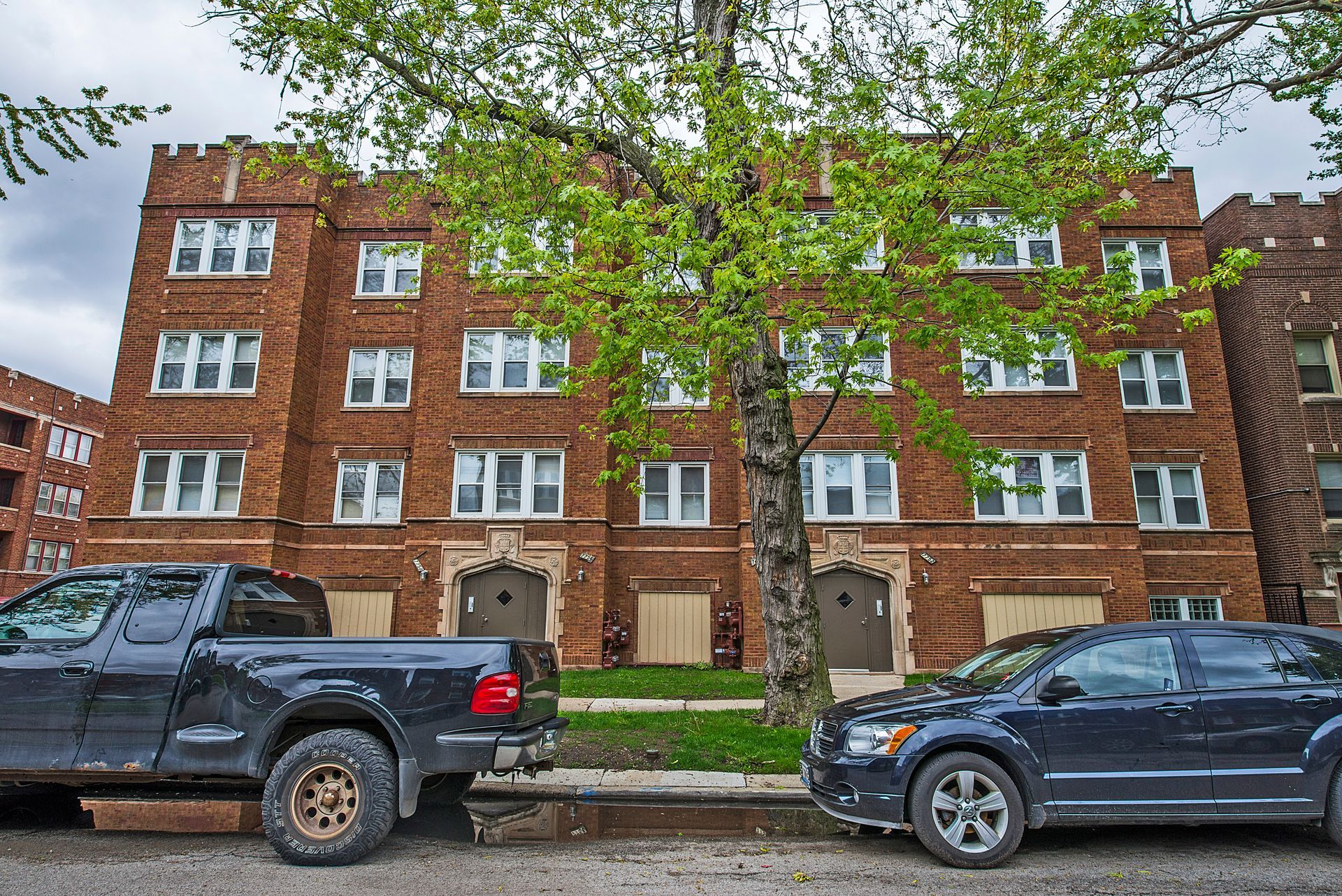 Brick apartment building with parked cars and a tree in front.