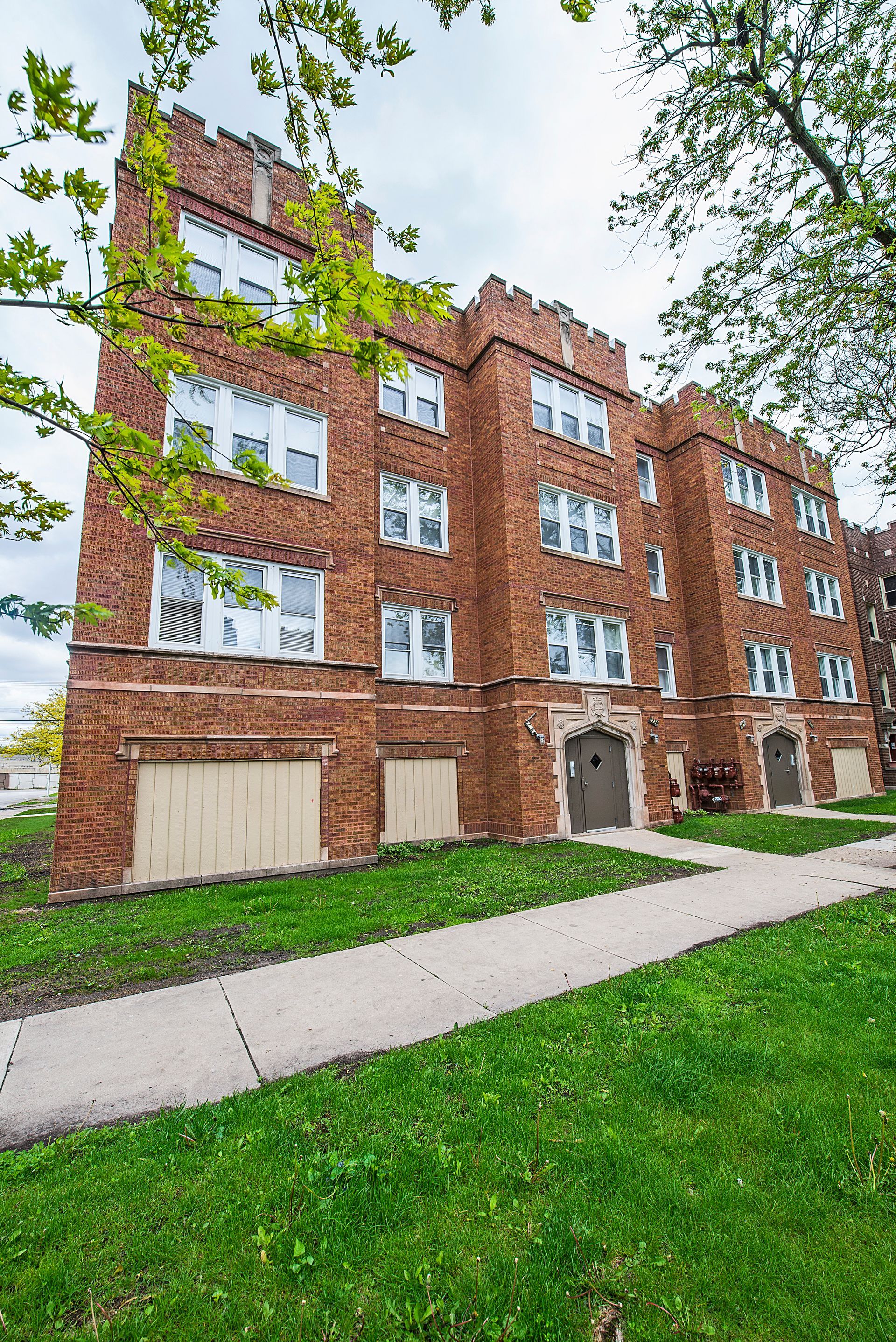 Brick apartment building with decorative crenellations, fronted by a sidewalk and grass.