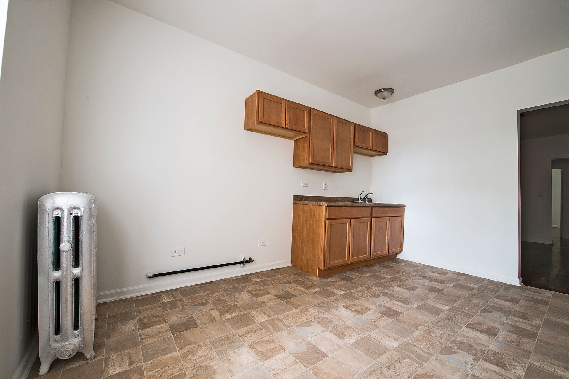 Small, empty kitchen with wood cabinets, radiator, and tiled floor.